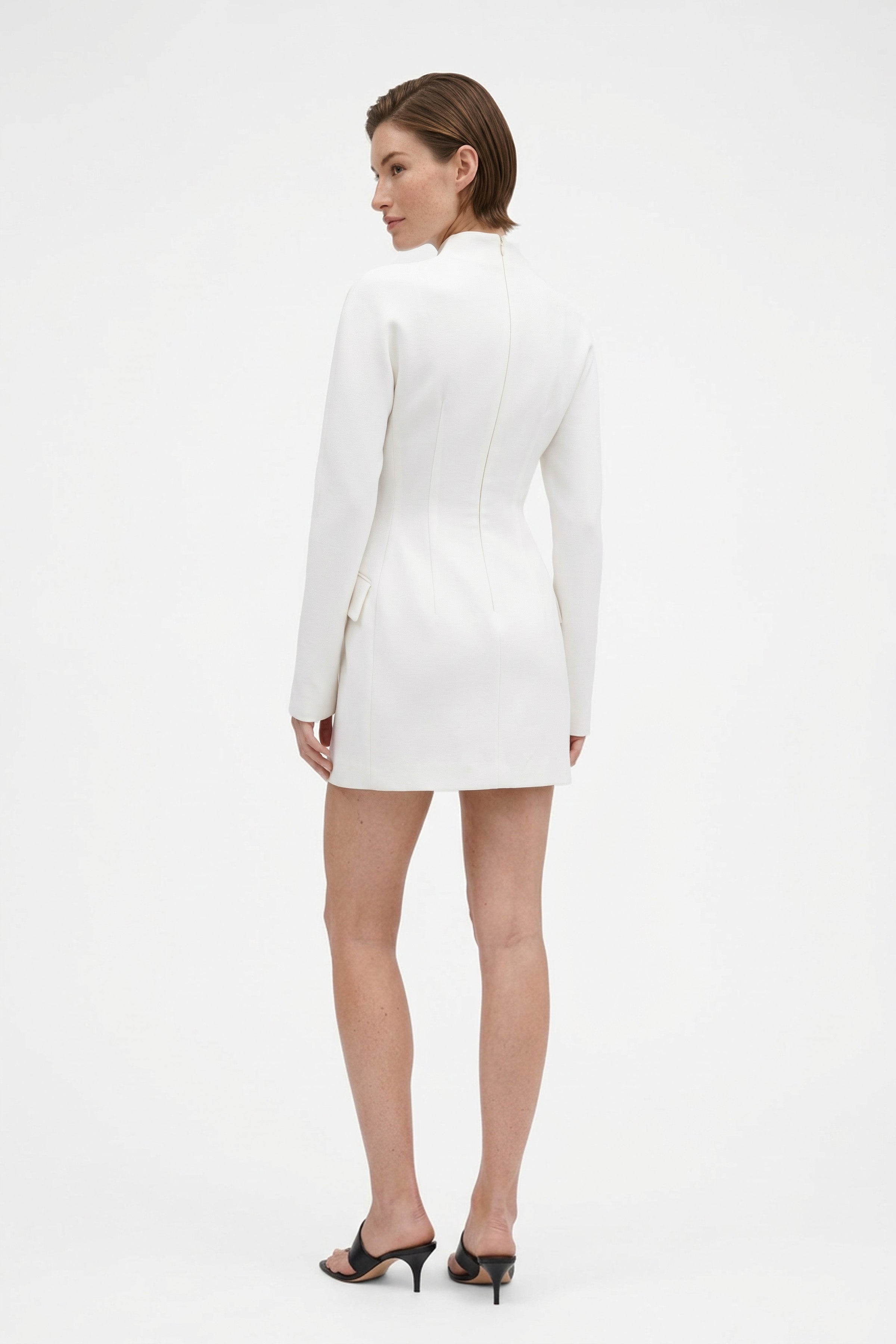 Model in white structured mini dress, back view, high neckline, long sleeves, heels.