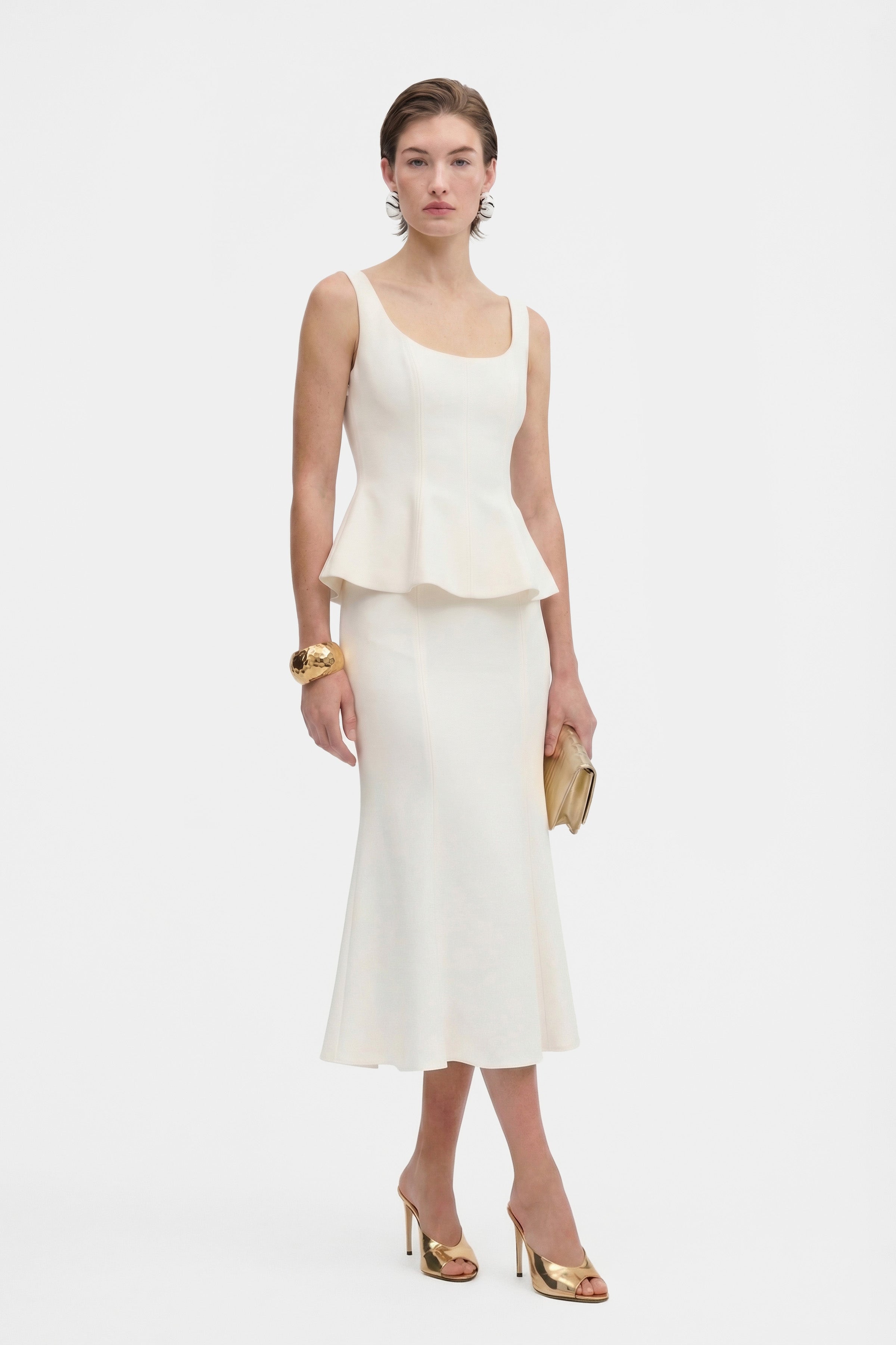 Model wearing ivory peplum top, front view with gold accessories, holding clutch.