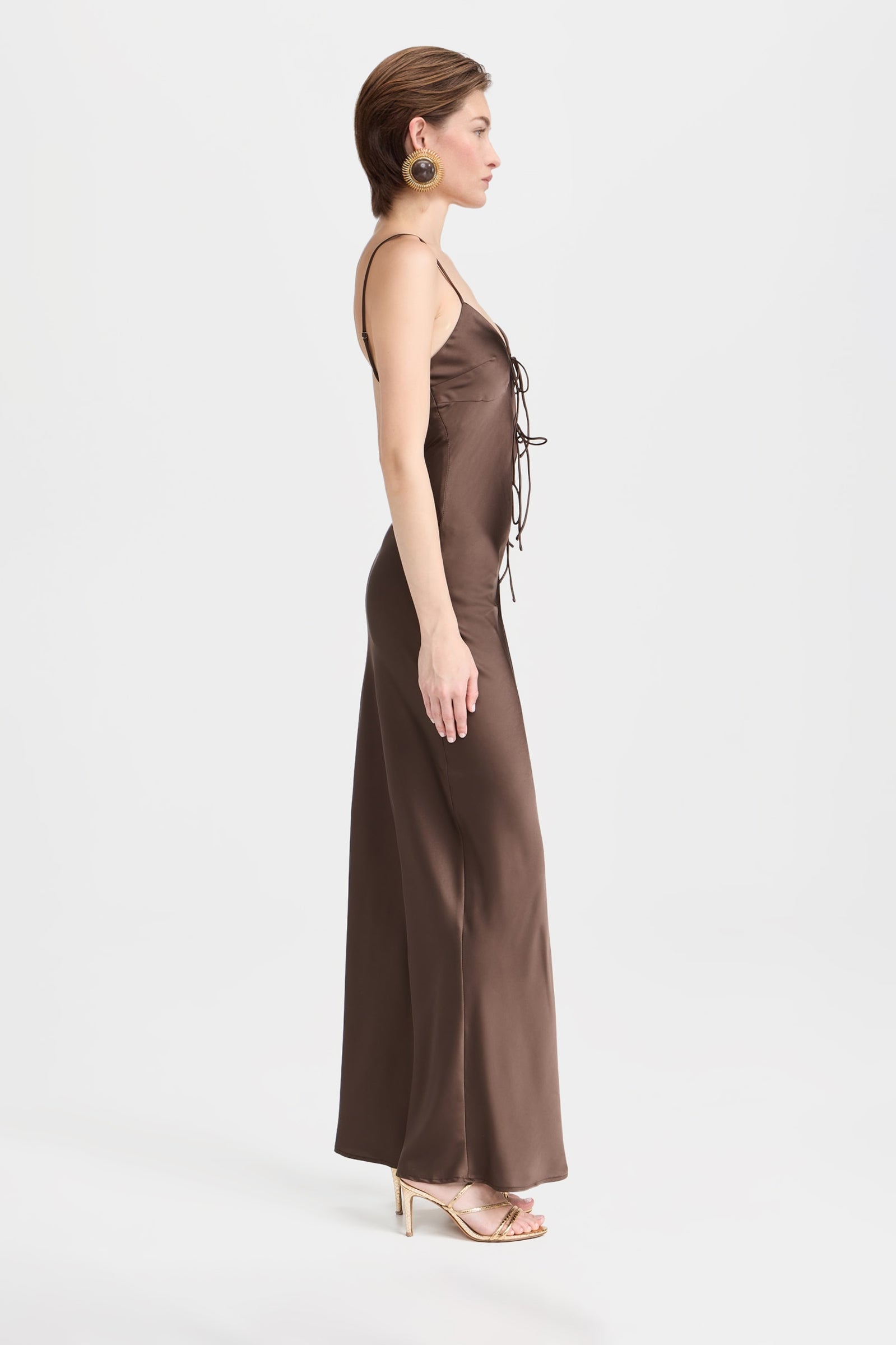 Full-length photo of a woman wearing an elegant dark brown satin slip dress, featuring a deep V-neckline, thin spaghetti straps, and unique side cut-outs with adjustable tie closures on the bodice.