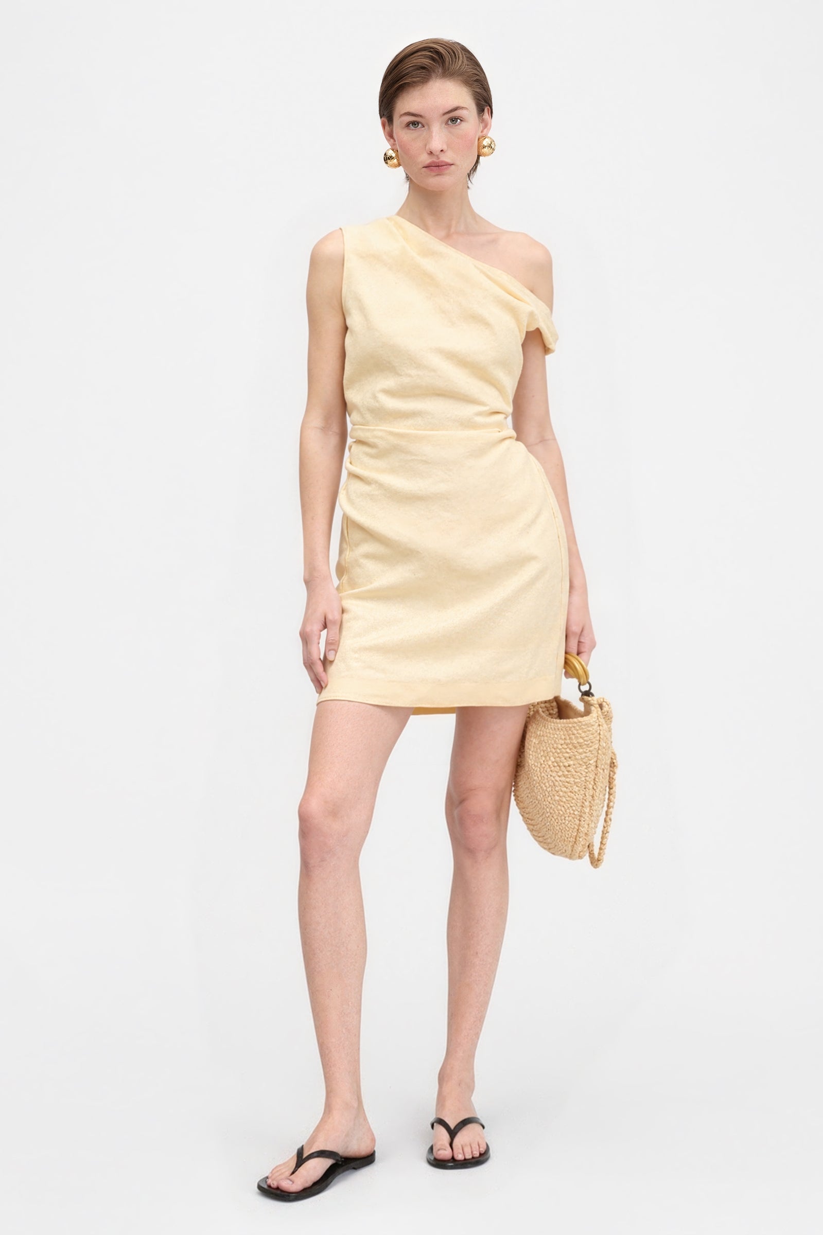 Model wearing a yellow linen mini dress, featuring a draped asymmetric one-shoulder design and a relaxed fit, paired with sandals.