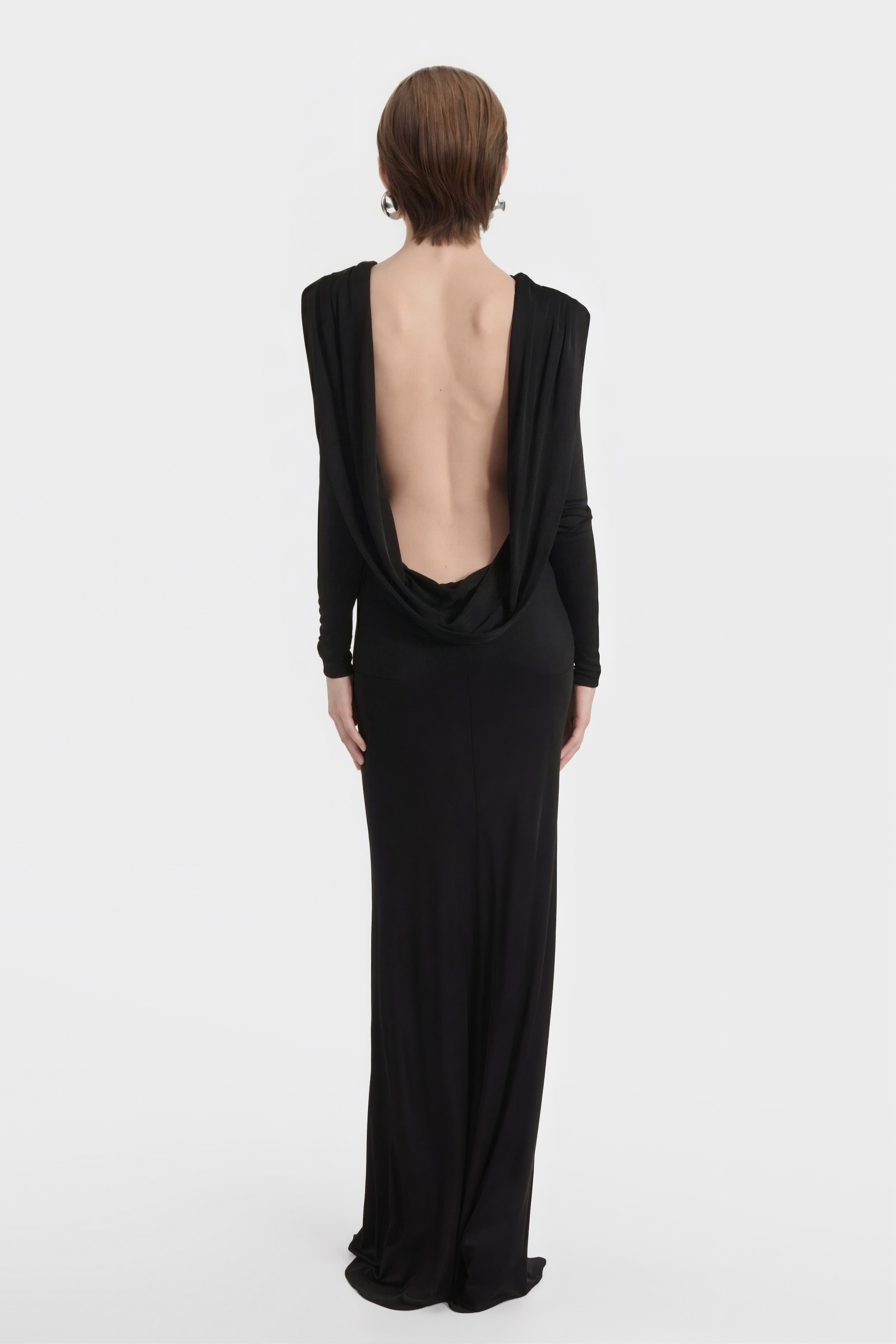 Full-length back view of a model showing the deep draped, open-back design of a black long-sleeved evening gown. The dress is floor-length and features a minimalist, sleek silhouette.
