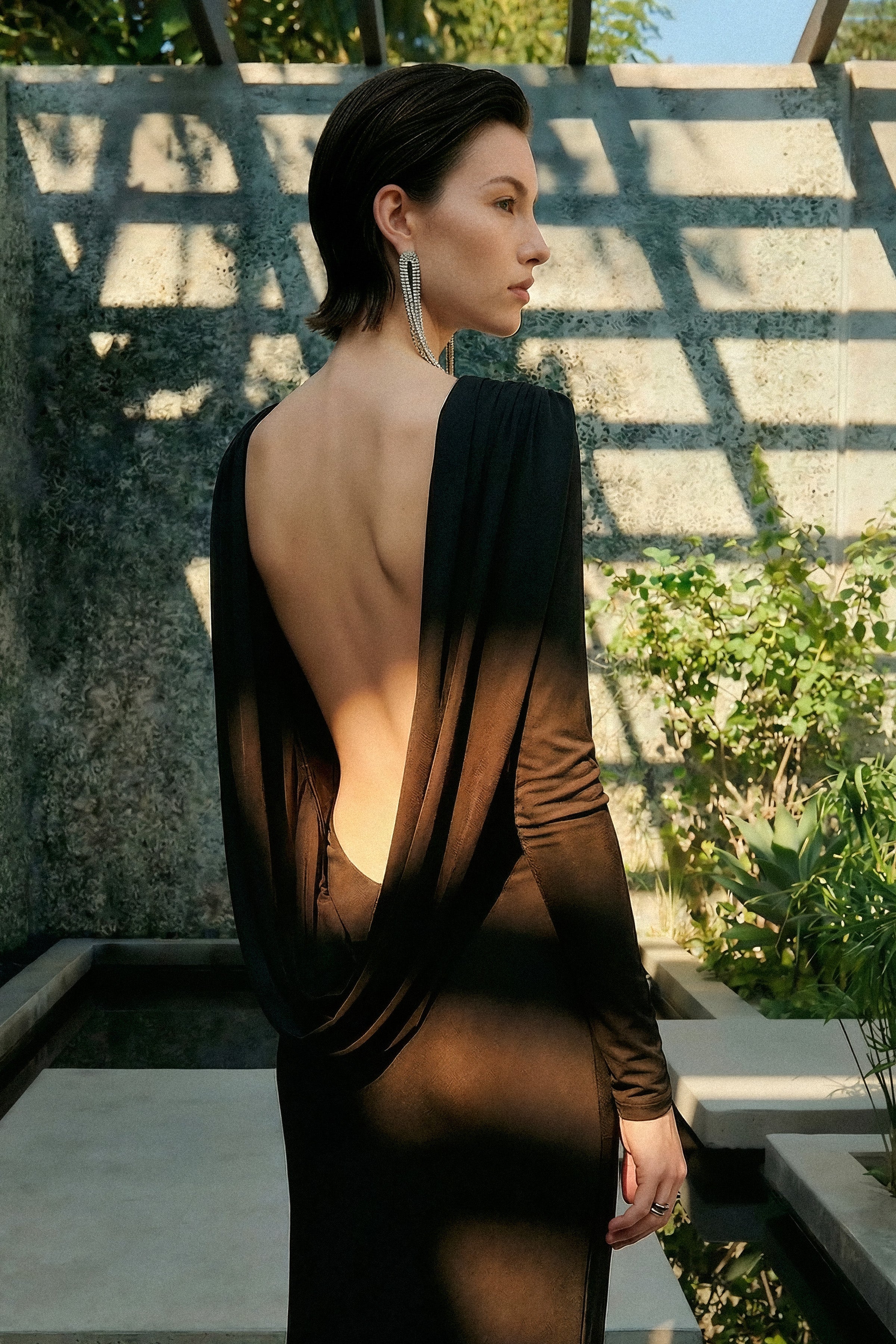 A close-up side profile and back view of a woman in a black backless gown featuring a deep draped cowl back. The shot is taken outdoors in natural light with shadows from a pergola falling across the model and a concrete wall.