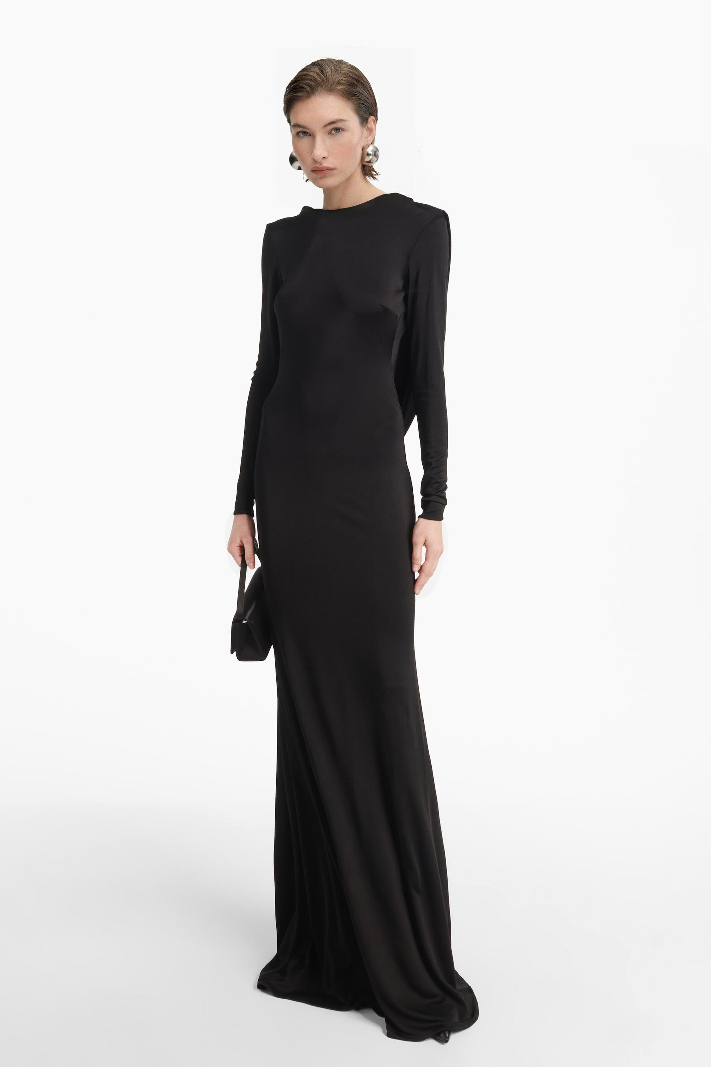 Full-length front view of a model wearing a floor-length black gown with long sleeves and a high neckline. She is holding a small black clutch and wearing large silver dome earrings against a bright white background.
