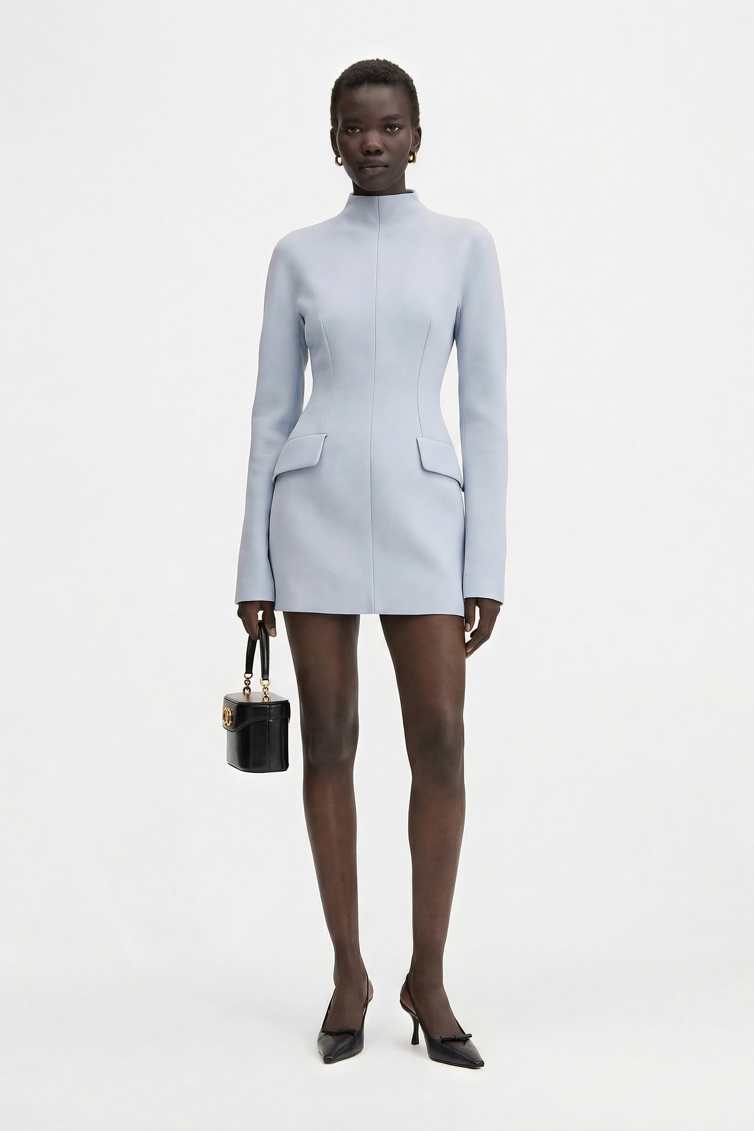 Full-length front view of a model wearing a structured long-sleeve light blue mini dress with a high neck, paired with black pointed-toe slingback heels and a small black leather handbag.