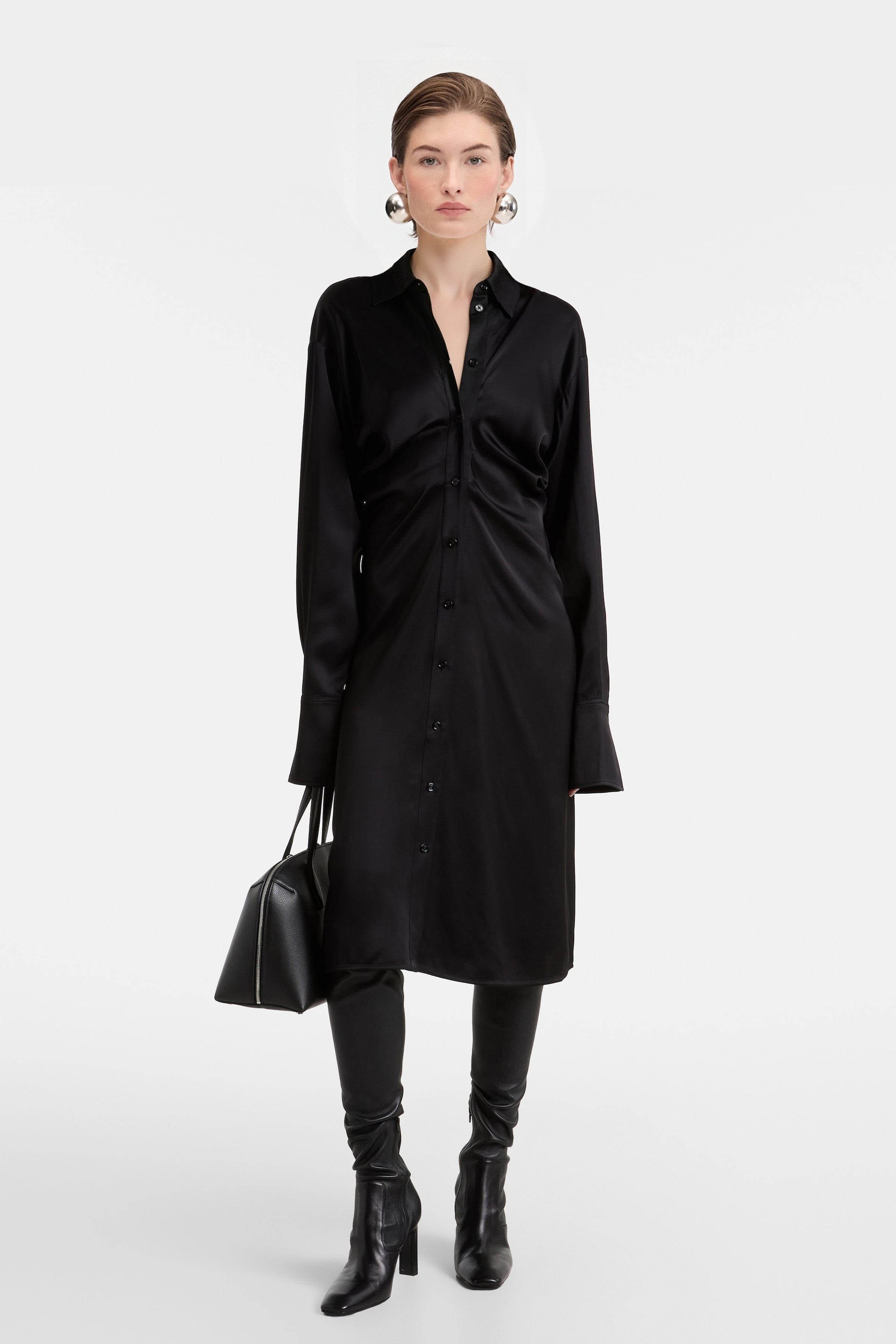 Full-body front view of a model wearing the Felipe Albernaz black satin midi shirt dress with ruched waist, styled with tall black leather boots and a matching handbag.