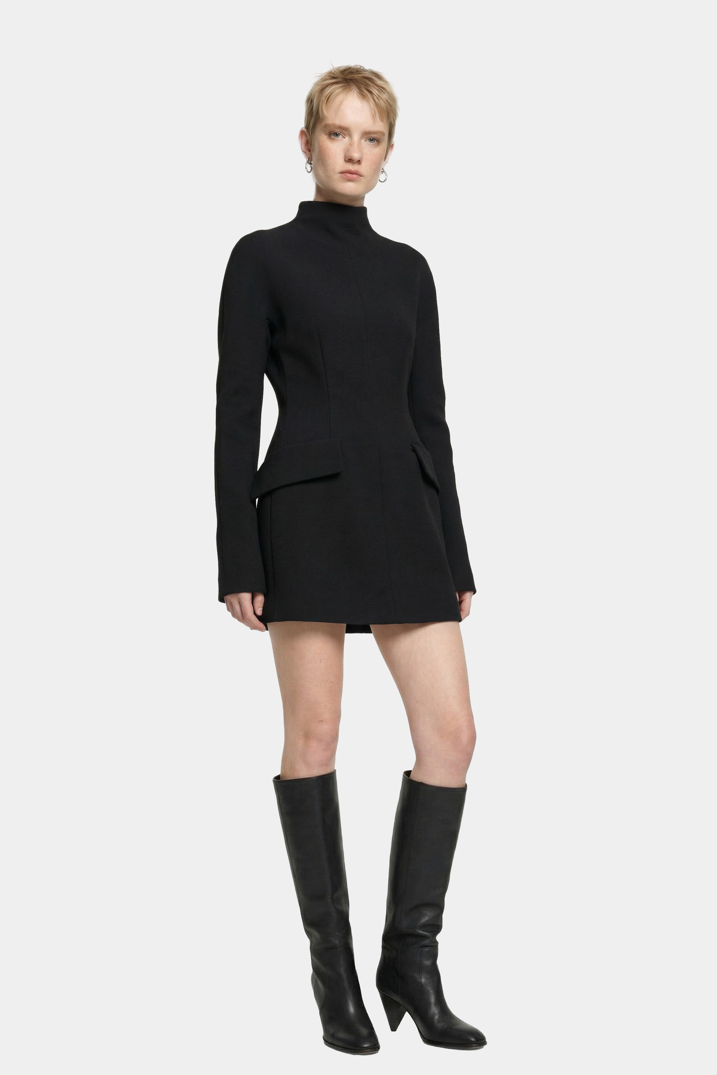 Model wearing black tailored mini dress, front view, paired with tall black boots.