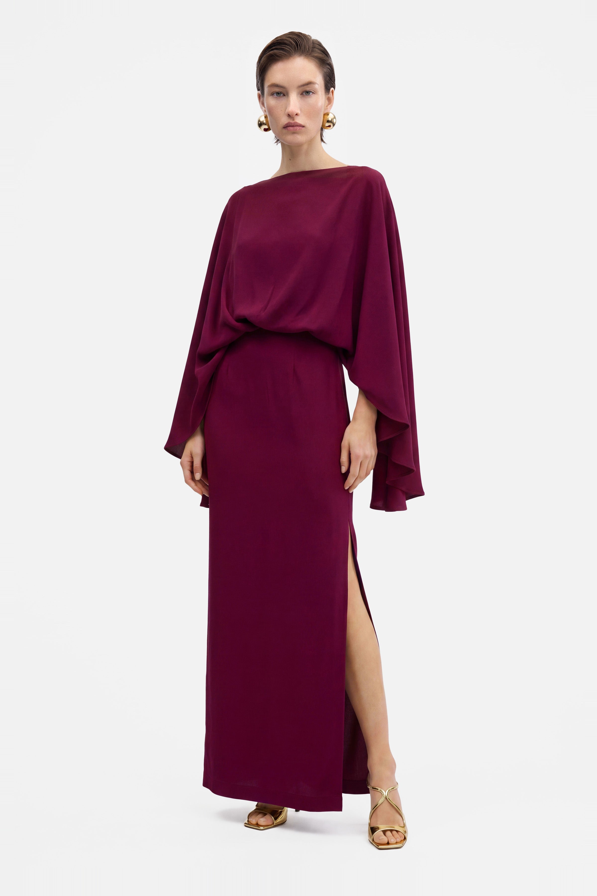 Model wearing a burgundy draped slit gown with flowing sleeves, front view.