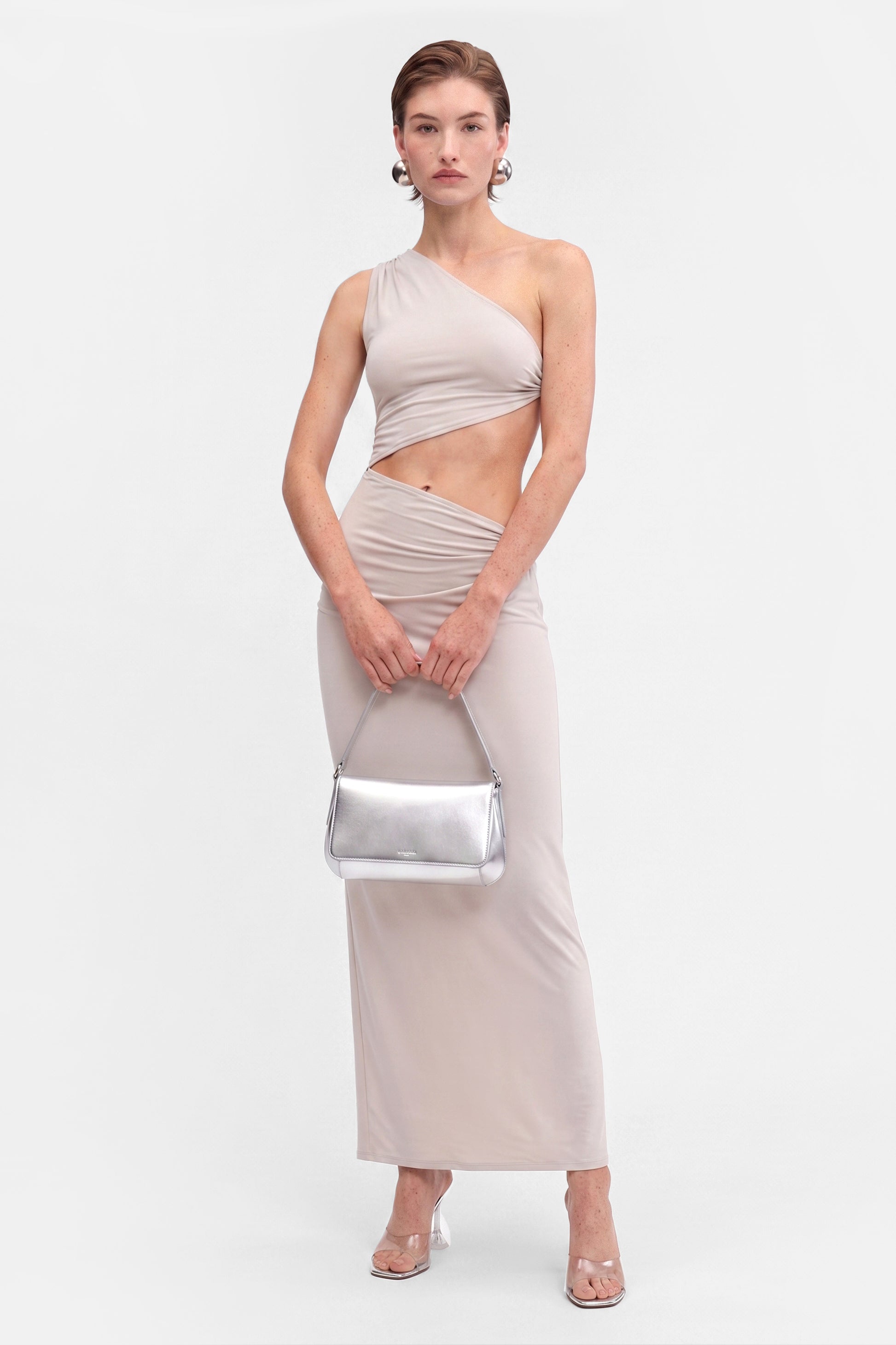 Model wearing asymmetric cutout maxi dress, holding silver bag, transparent heels. Front view.