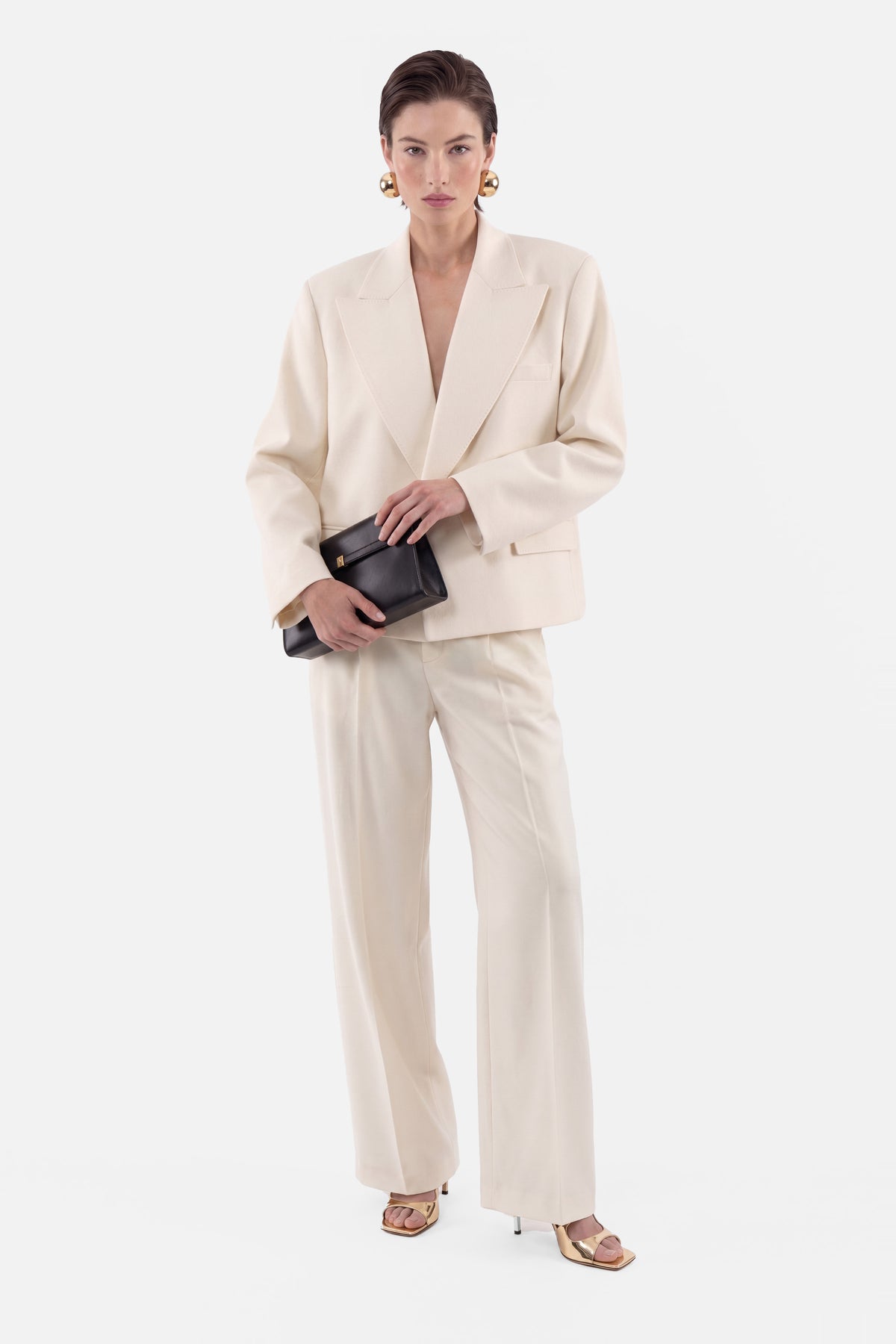 Ivory tailored suit, front view, model holding black clutch, wearing gold earrings.
