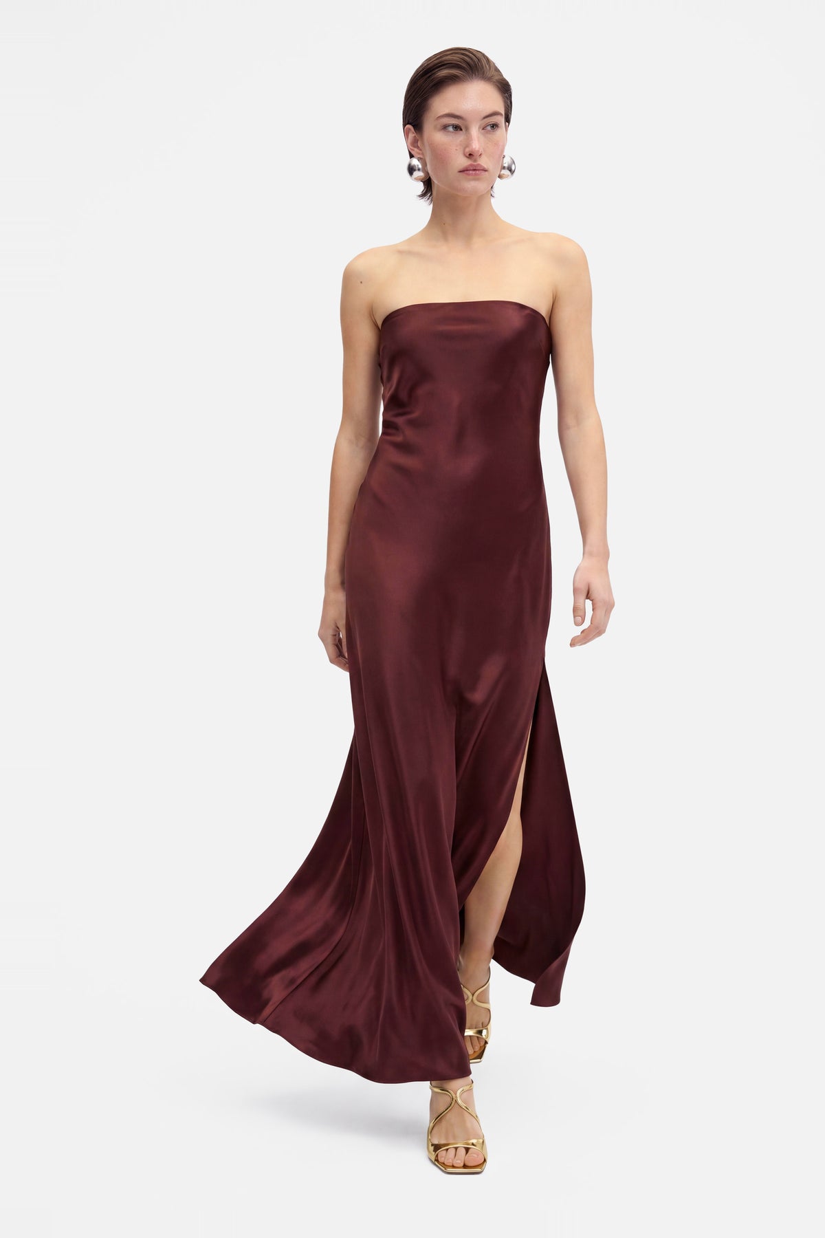 Model wearing burgundy strapless gown, thigh-high slit, flowing front view, gold sandals.
