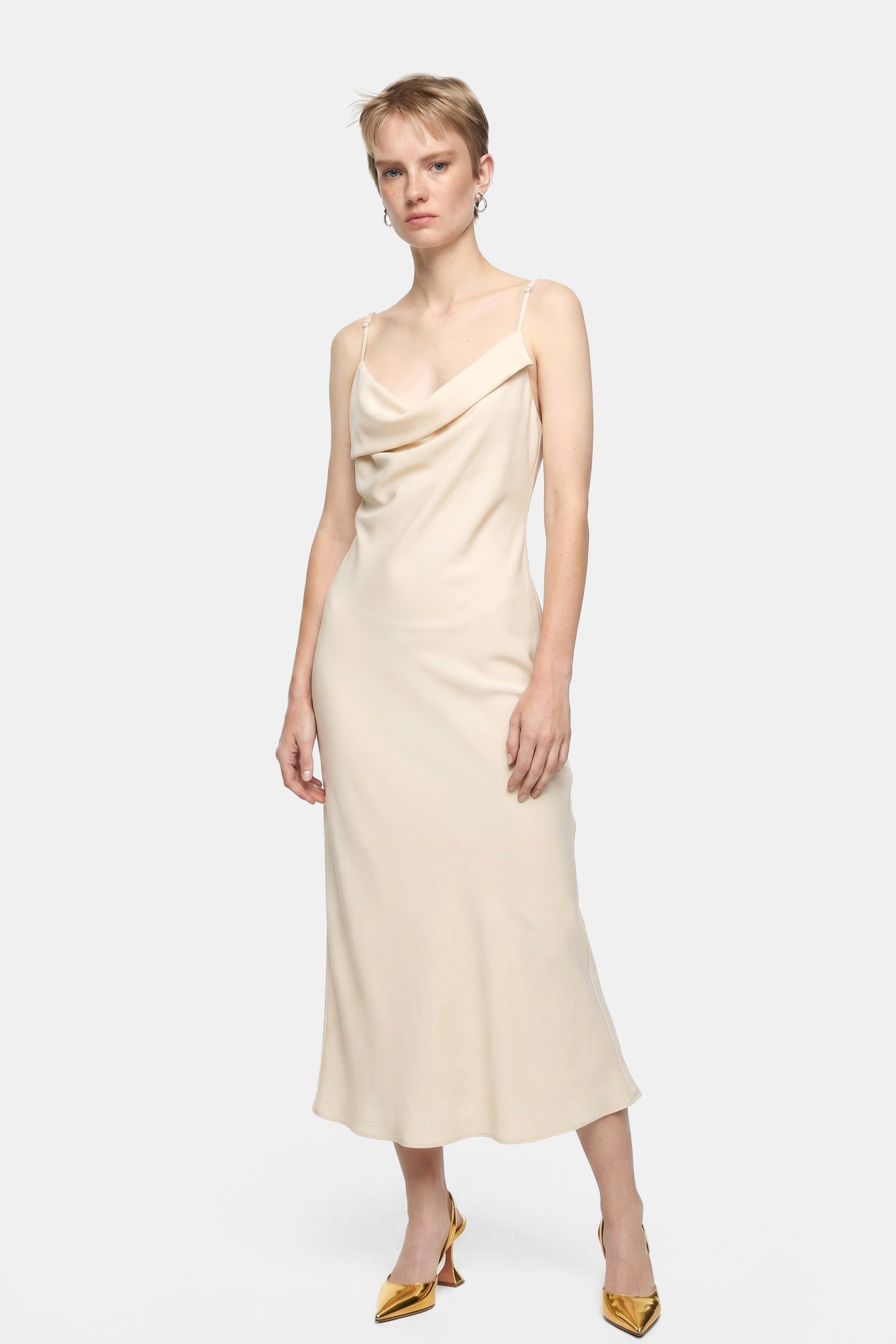Model wearing a cream silk cowl neck slip dress, gold heels; front view.