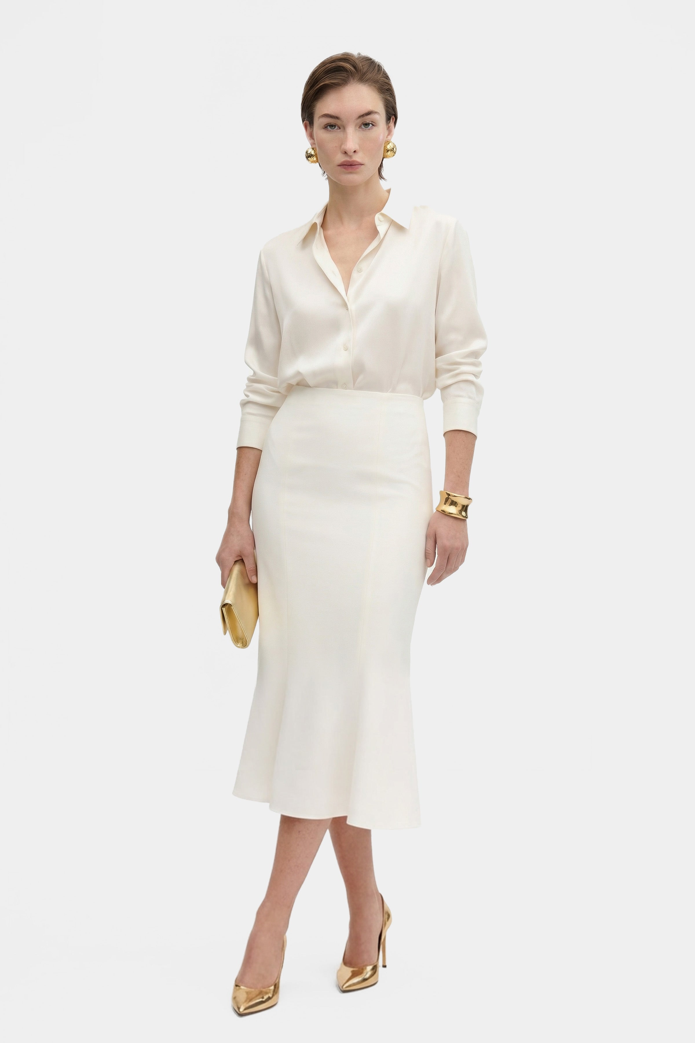 Front view of model in elegant ivory midi skirt, gold accessories, holding clutch.