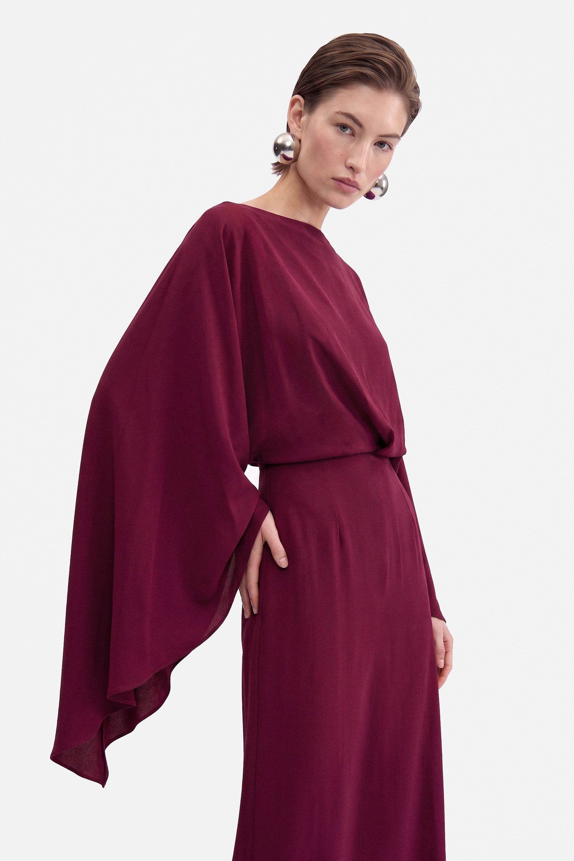 Front view of burgundy draped slit gown, elegant flowy design with cape sleeves.