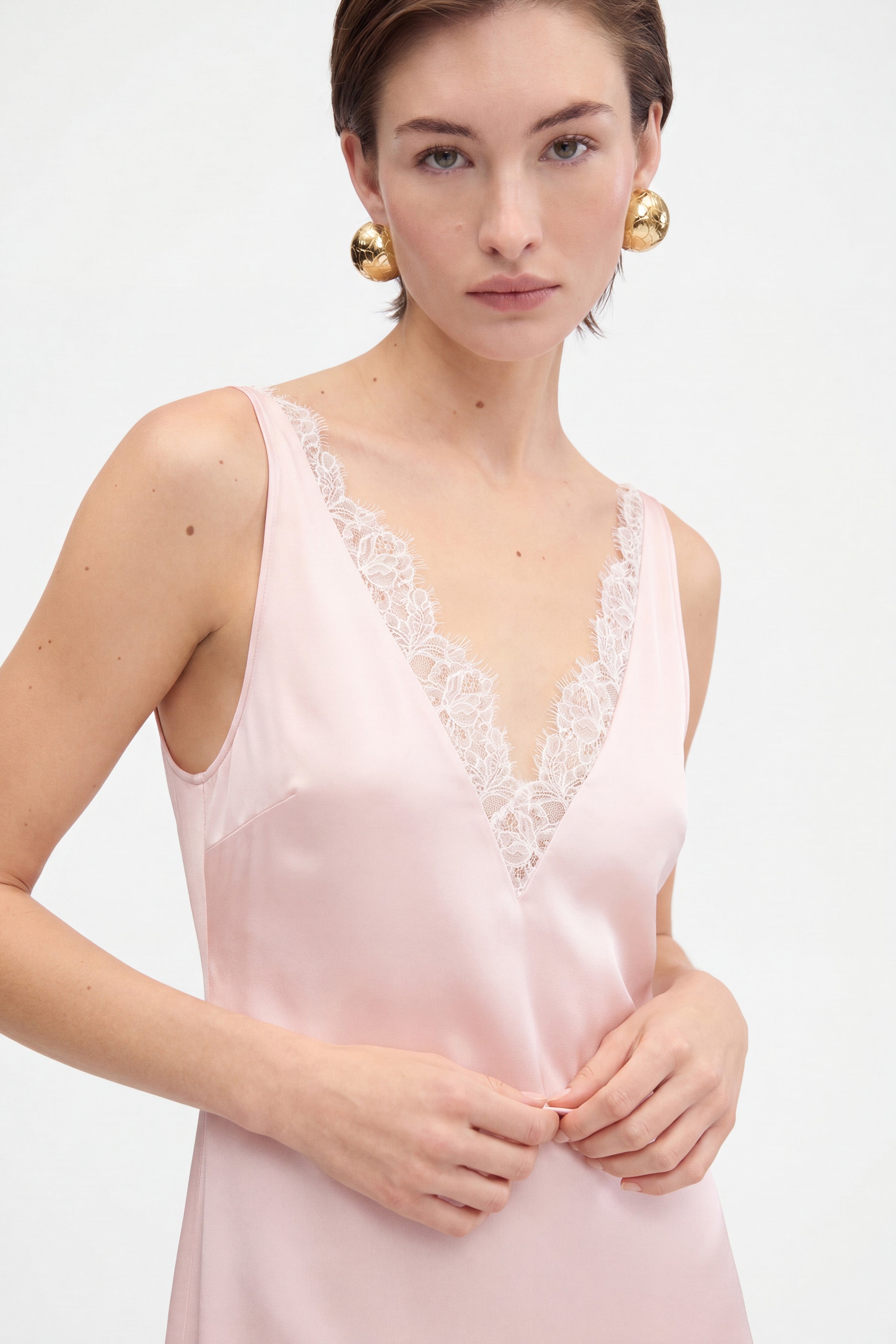 Blush lace slip dress front view, lace detail, model with gold earrings, pale background.