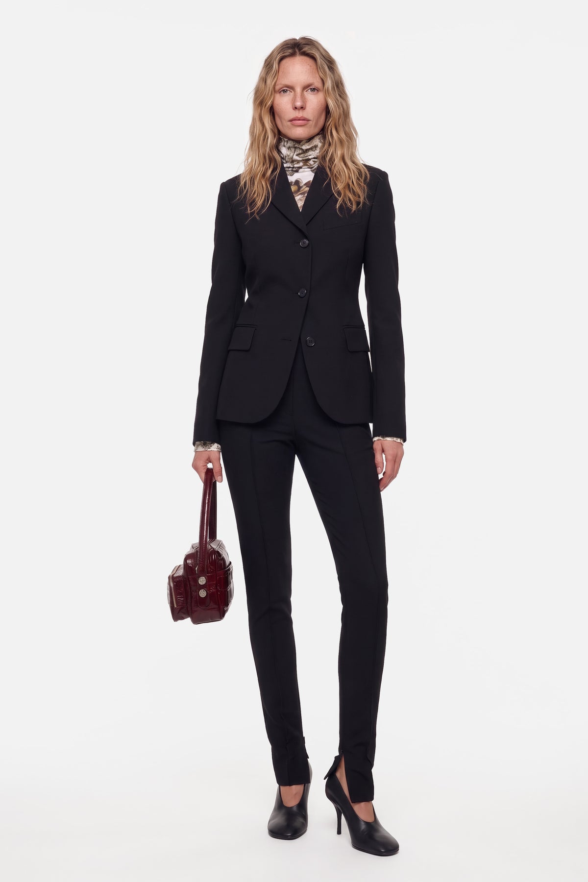 Model in tailored black suit, front view, holding brown handbag, wearing heels.