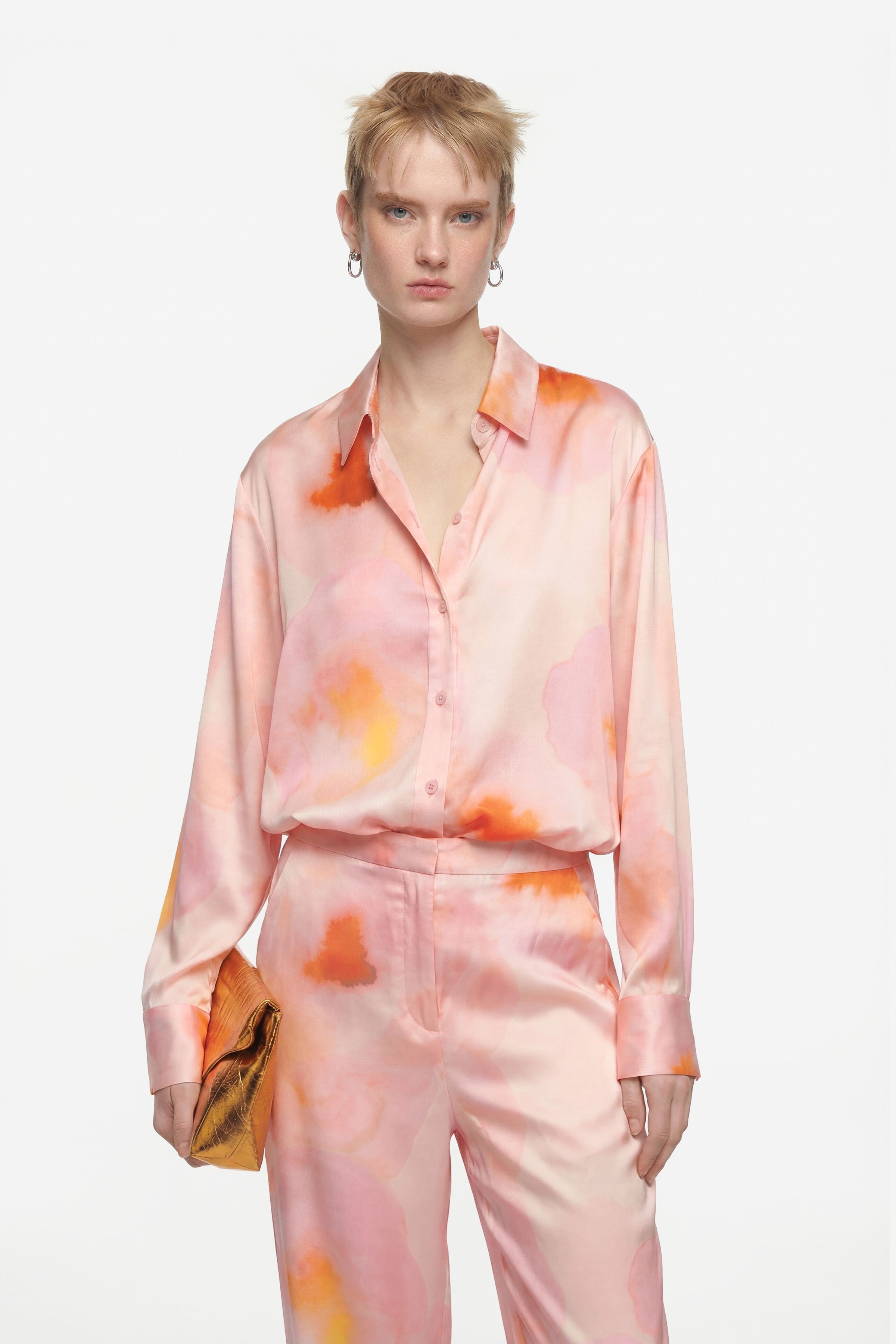 Model wearing pink-orange watercolor silk set, holding gold clutch. Front view, long sleeves.