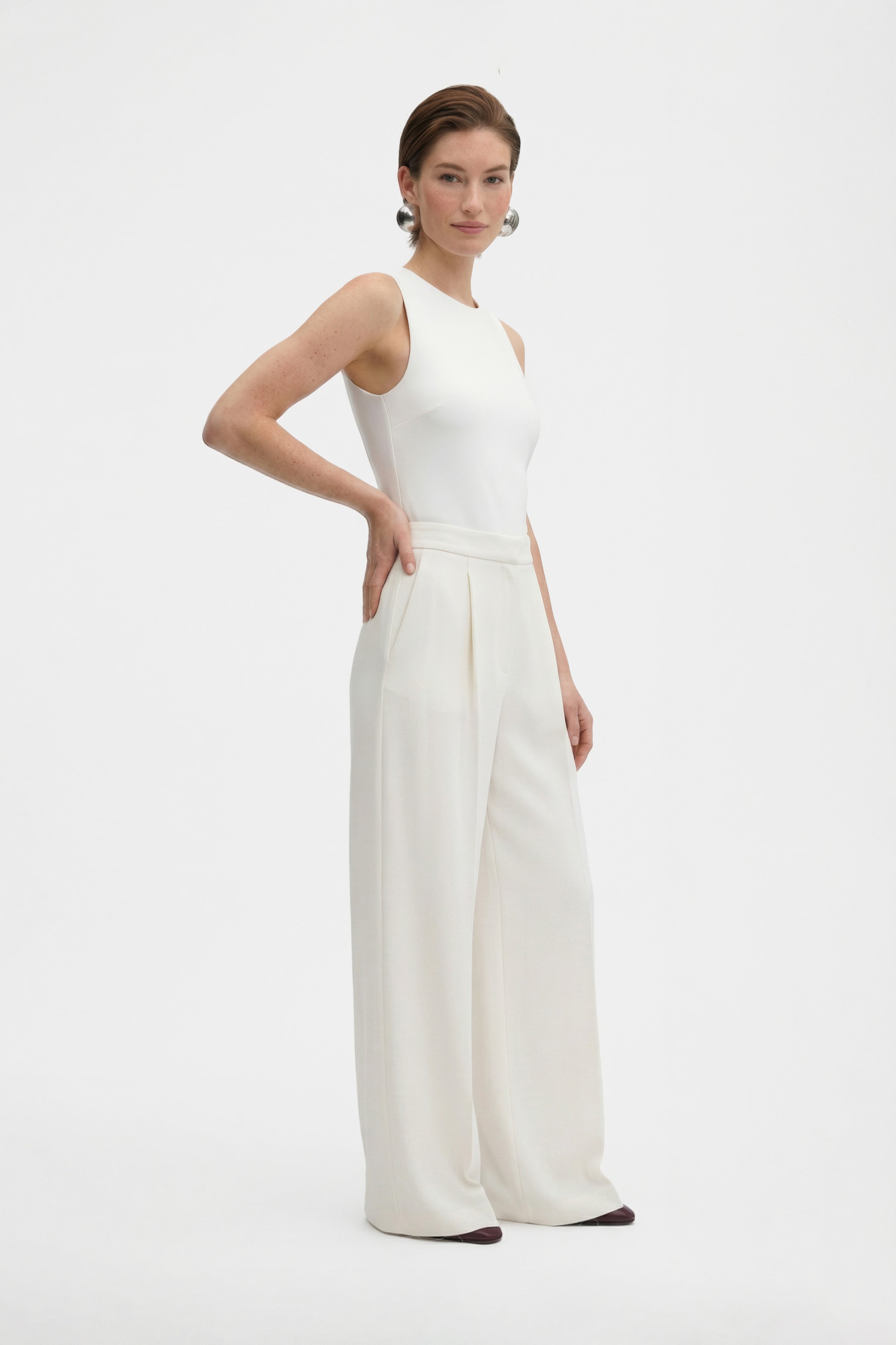 Model wearing ivory wide-leg trousers, side view, with hands on waist, neutral background.