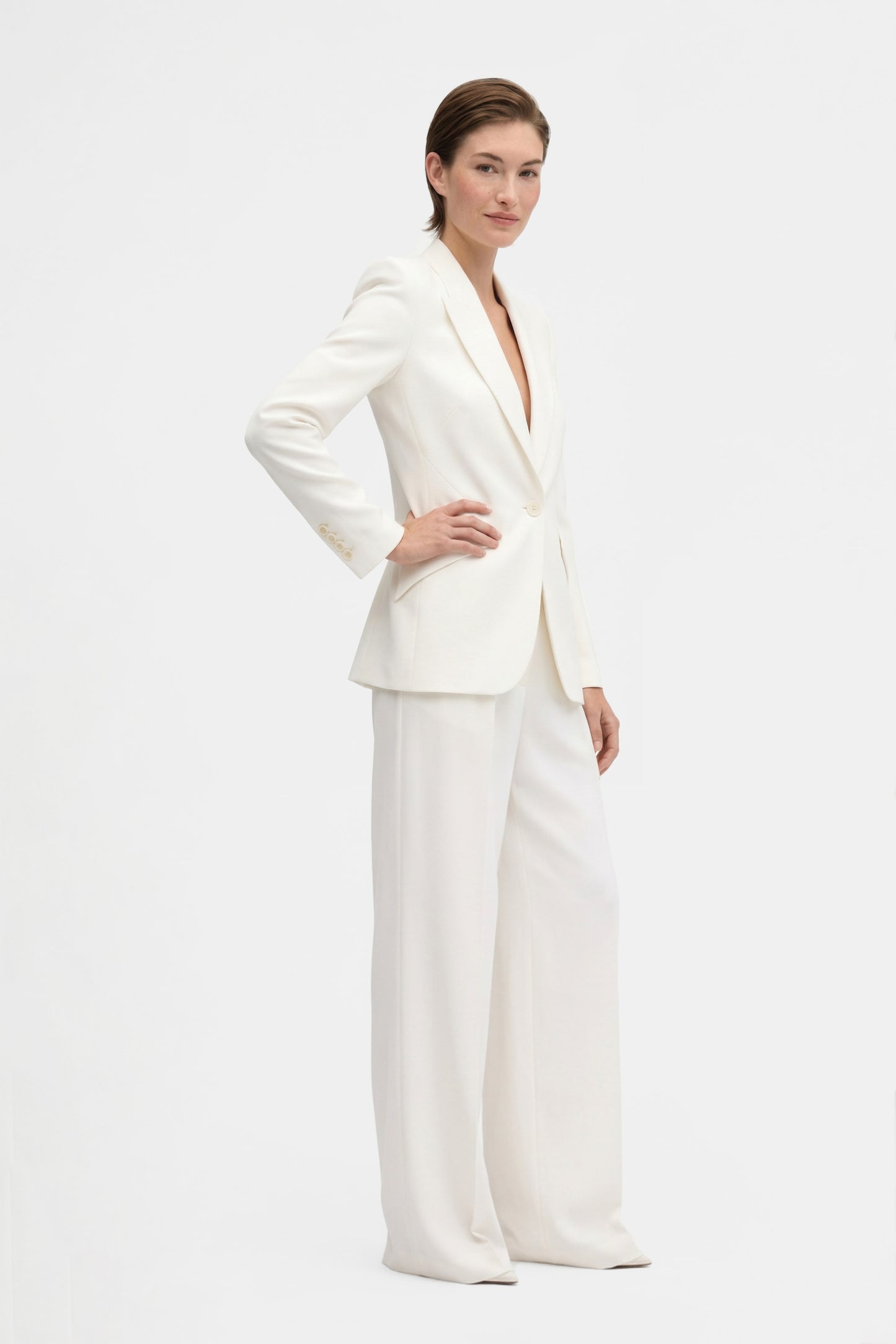 Model wearing ivory tailored blazer, side view, hands on hips, minimal background.