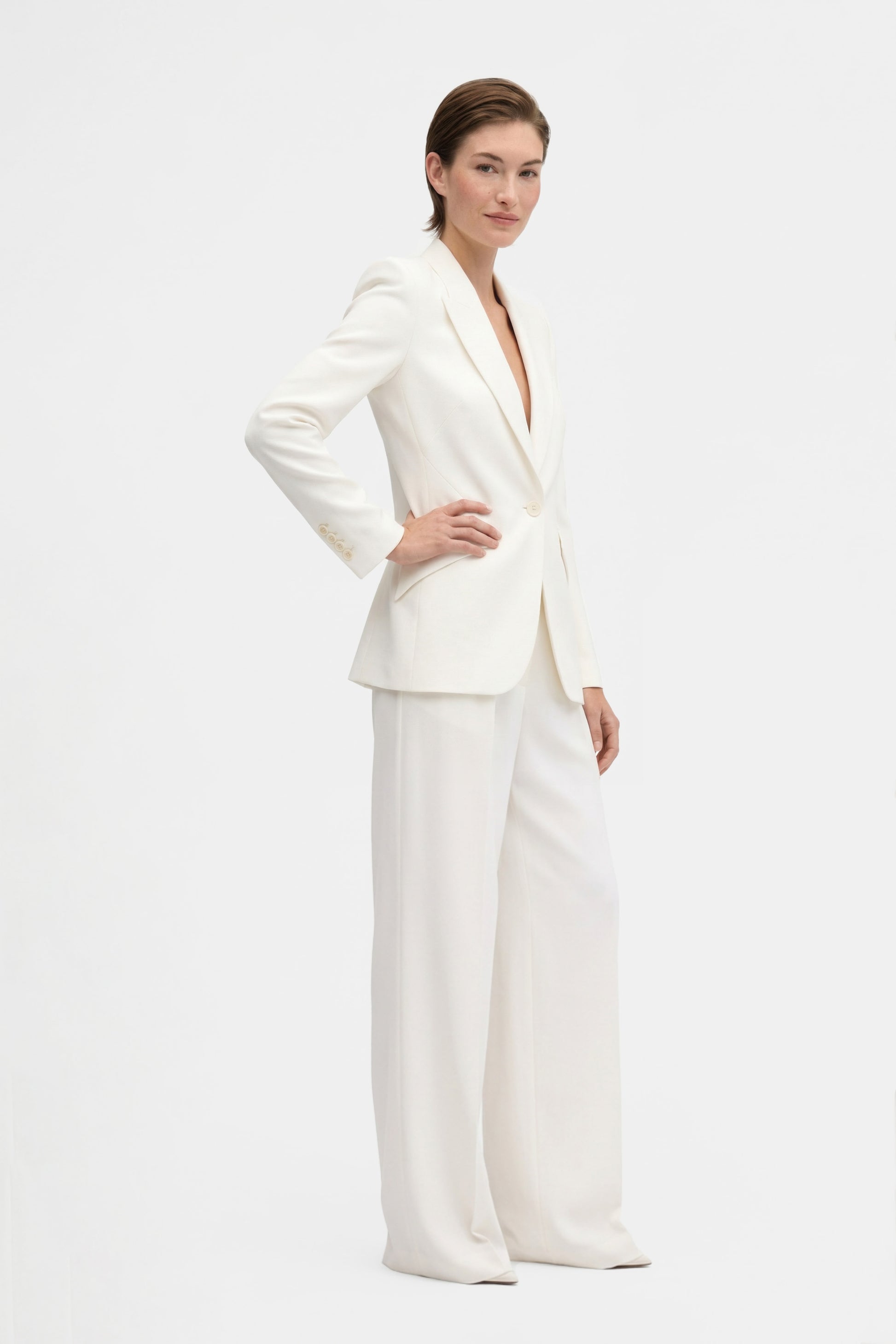 Model wearing ivory tailored blazer, side view, hands on hips, minimal background.