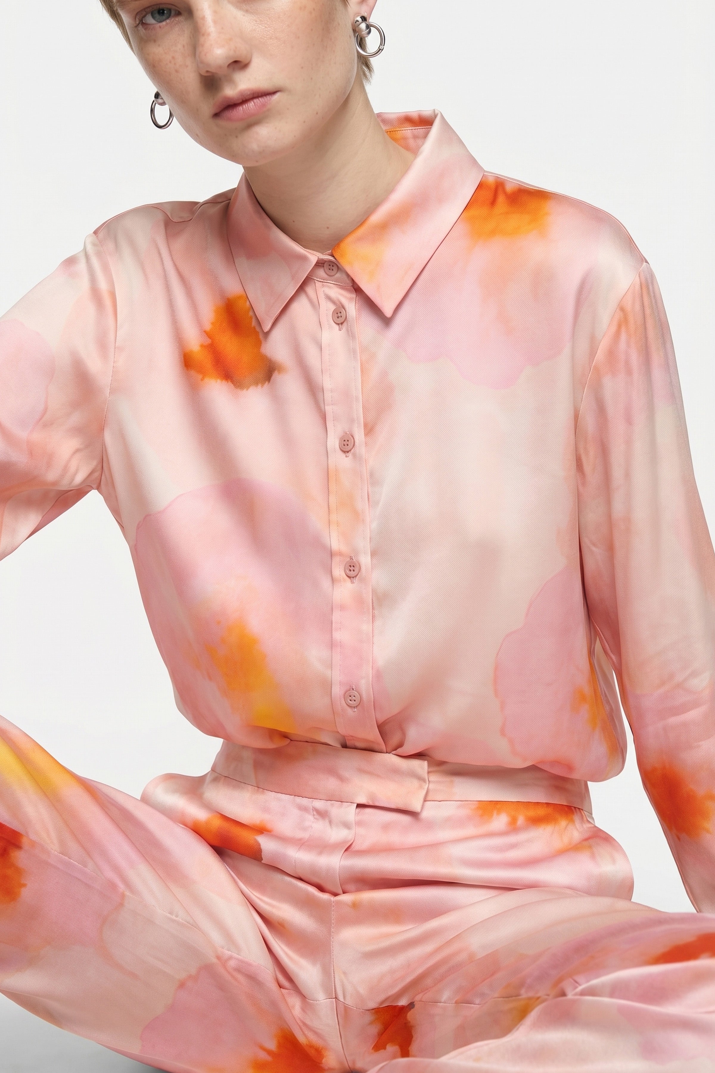 Model seated in pink and orange watercolor-patterned silk blouse and pants, front view.