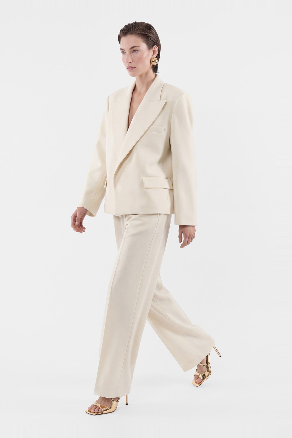 Model walking in an ivory tailored suit, front view, gold heels.
