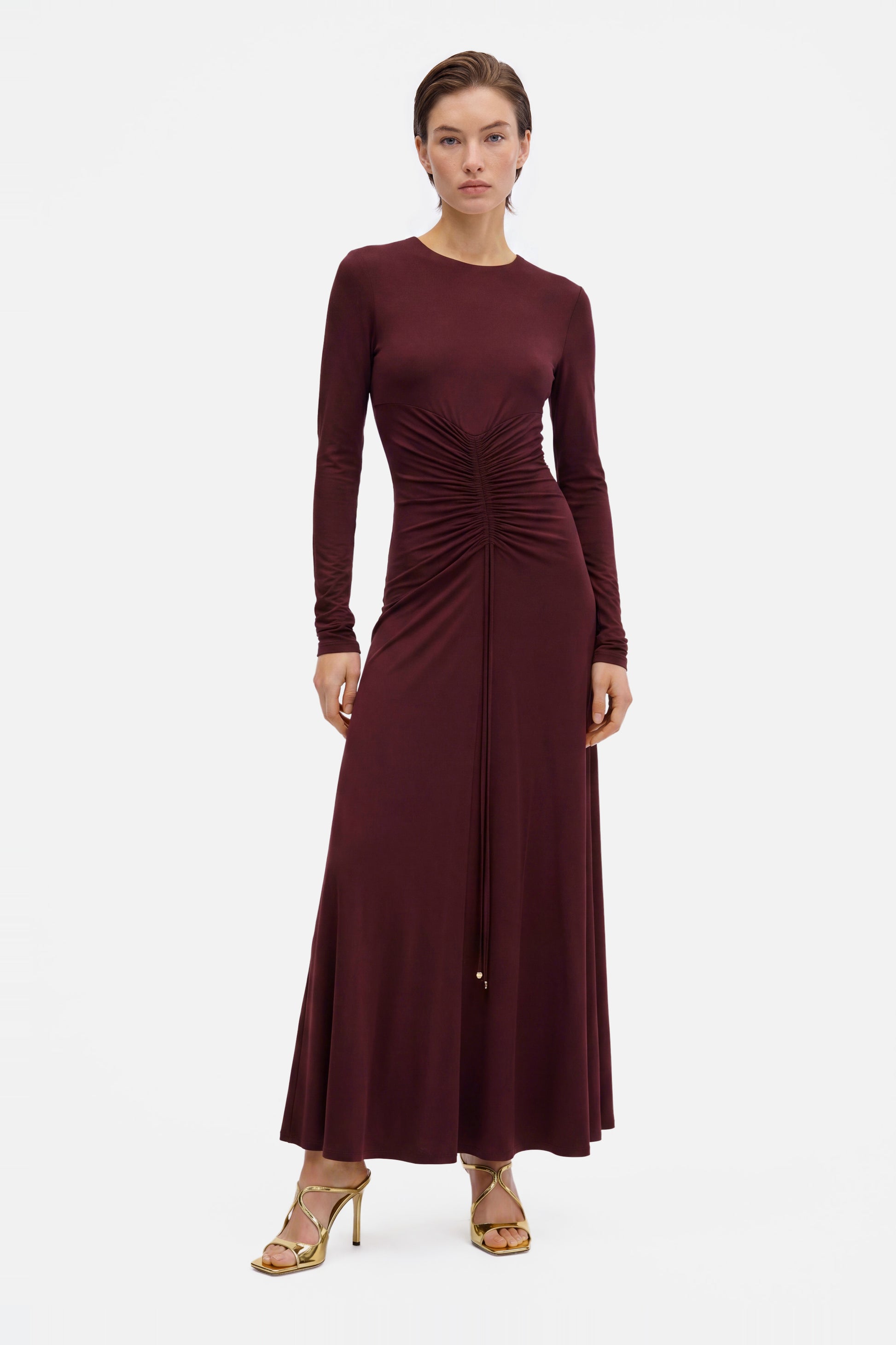 Front view: Burgundy ruched long sleeve dress, model standing, gold heels.