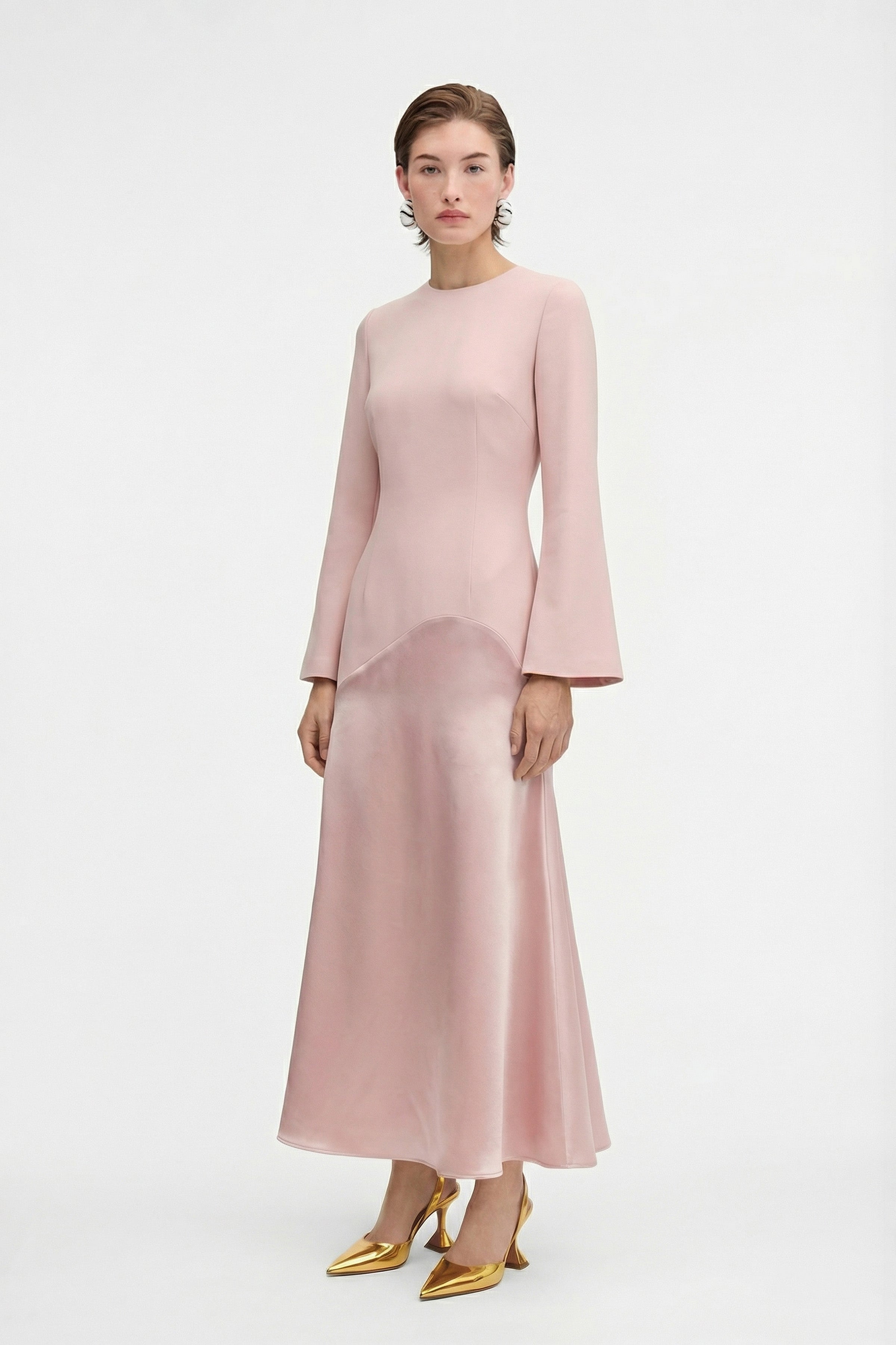 Model standing in a pink satin-crepe blend long dress with flared hem and long sleeves, holding a gold pouch bag.