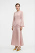 Model standing in a pink satin-crepe blend long dress with flared hem and long sleeves, holding a gold pouch bag.