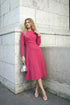 Model wearing elegant fuchsia midi dress, front view, leaning against stone wall.