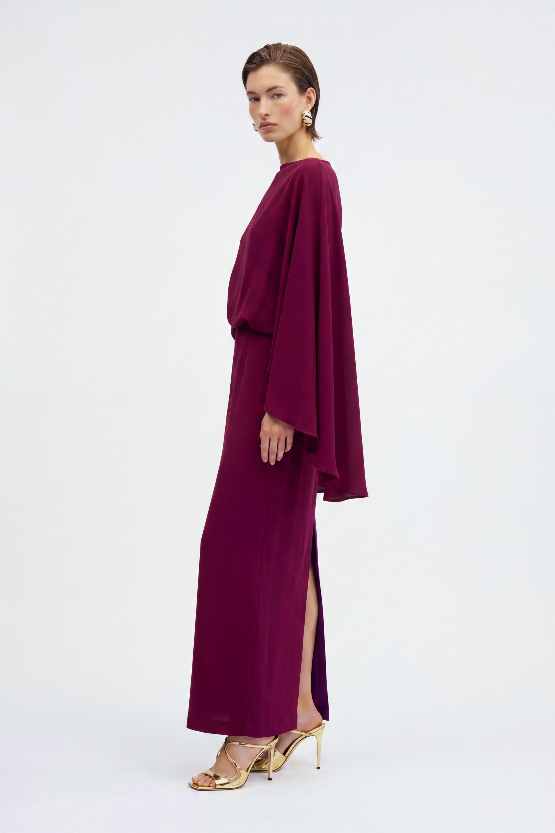 Model in burgundy draped slit gown, side view, gold heels, minimalist backdrop.