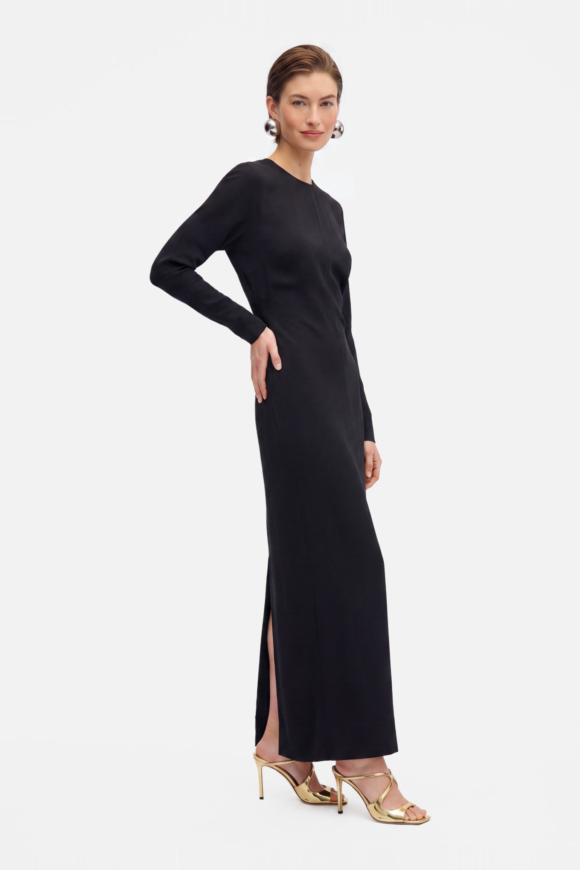 Model wearing black long sleeve maxi dress, side slit, gold heels, elegant look.