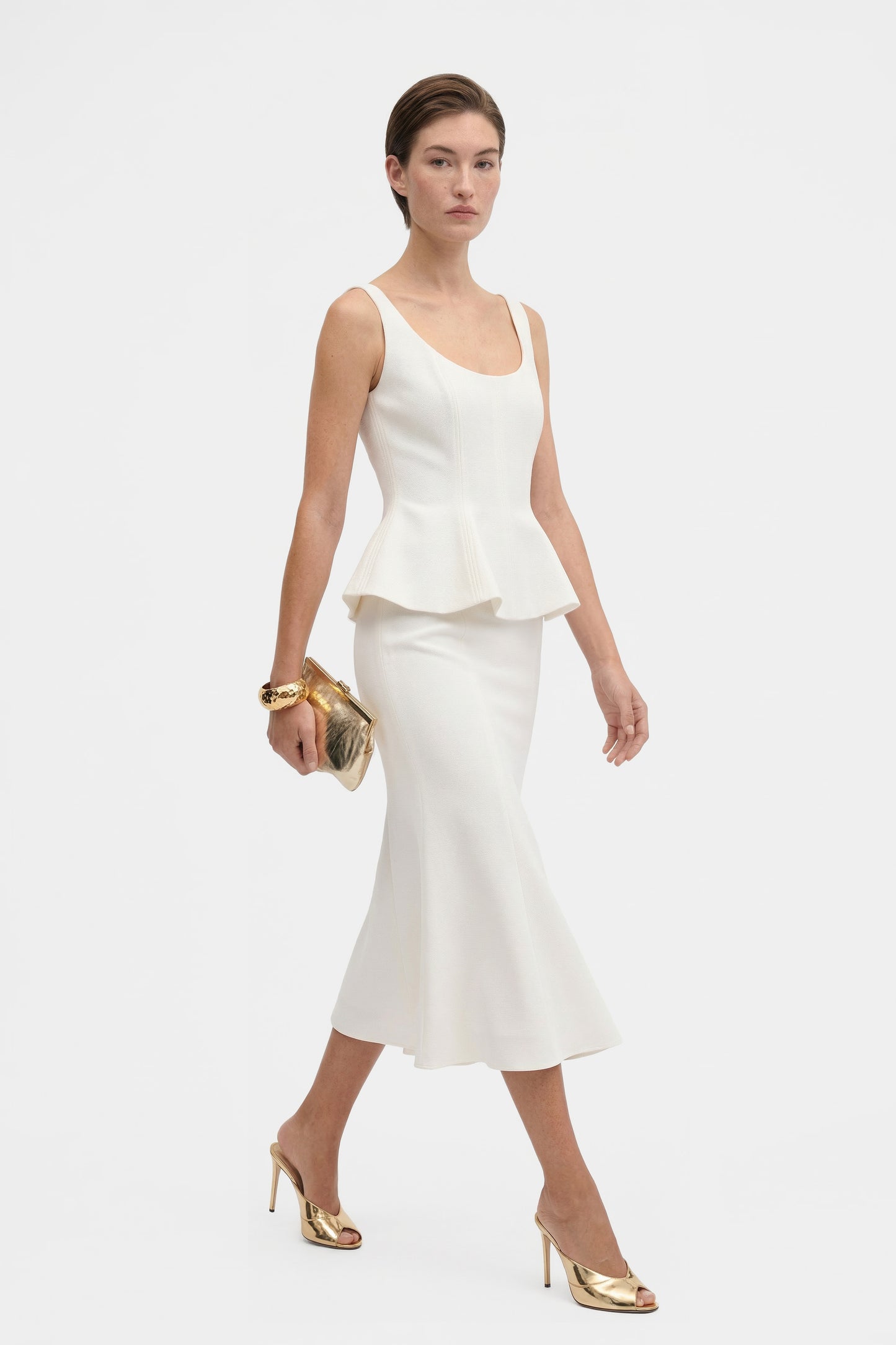 Model walking in ivory peplum top, gold shoes, front view. Elegant styling.