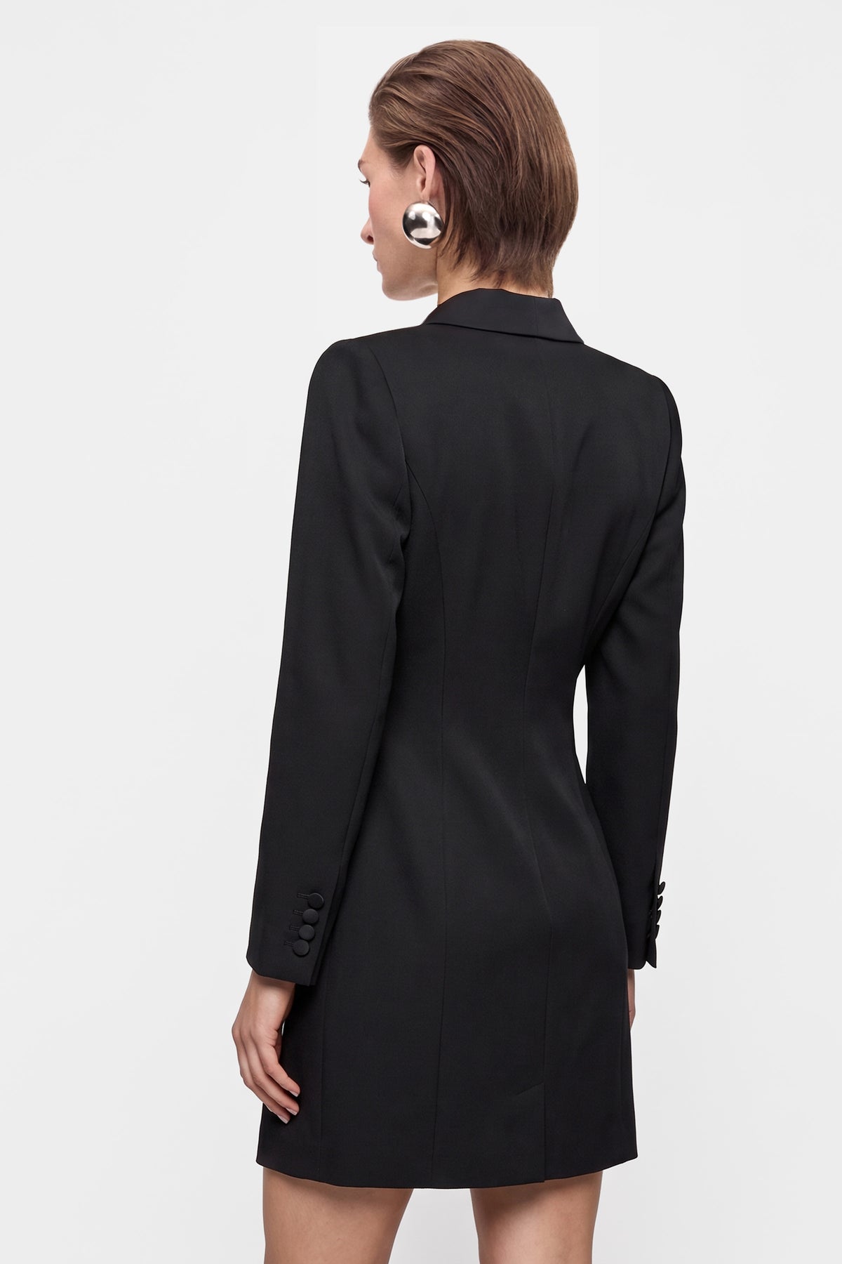 Model wearing black tuxedo mini dress, back view, fitted silhouette, buttoned sleeves.