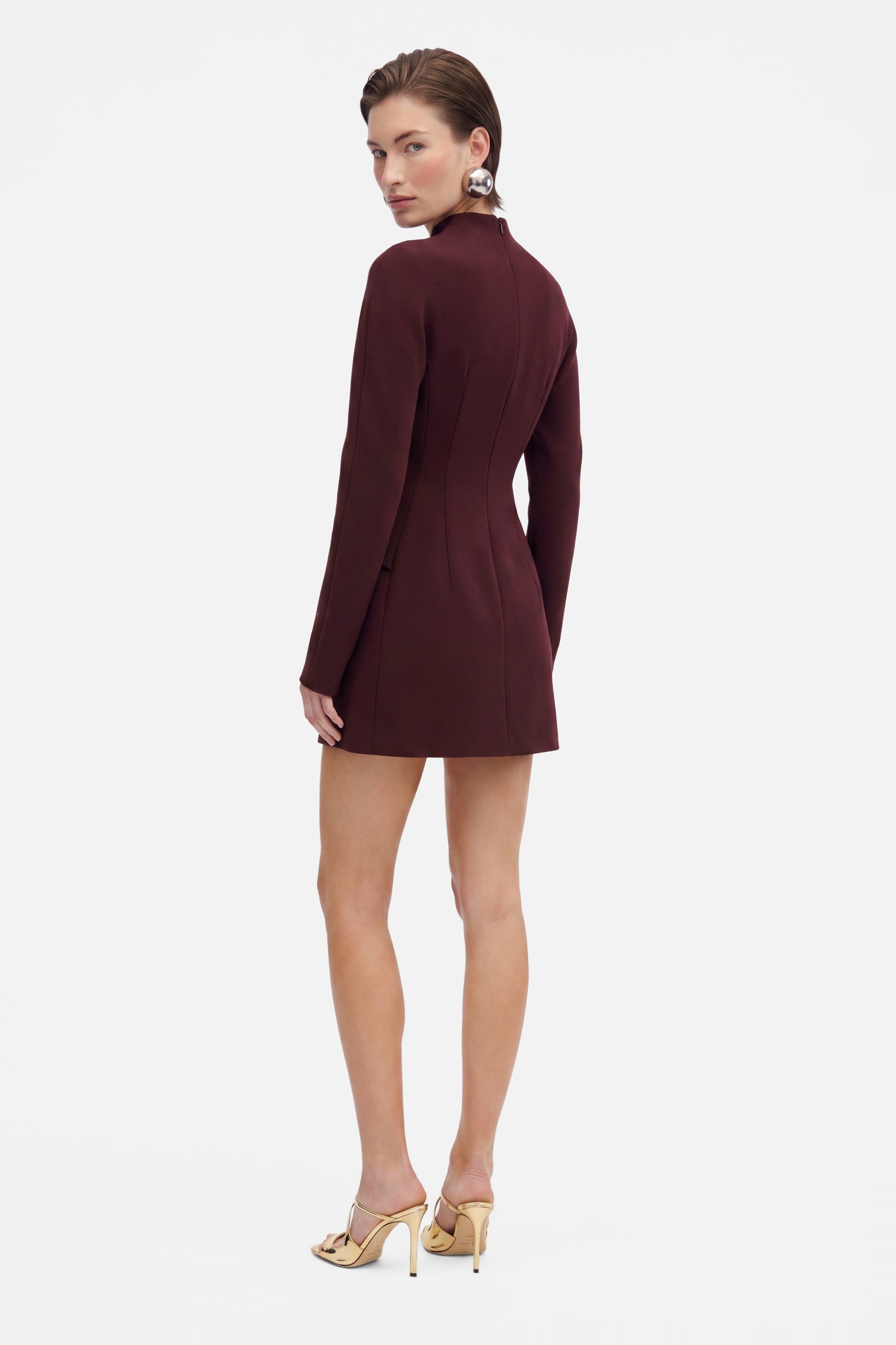 Back view of a model in a burgundy structured mini dress, gold heels.