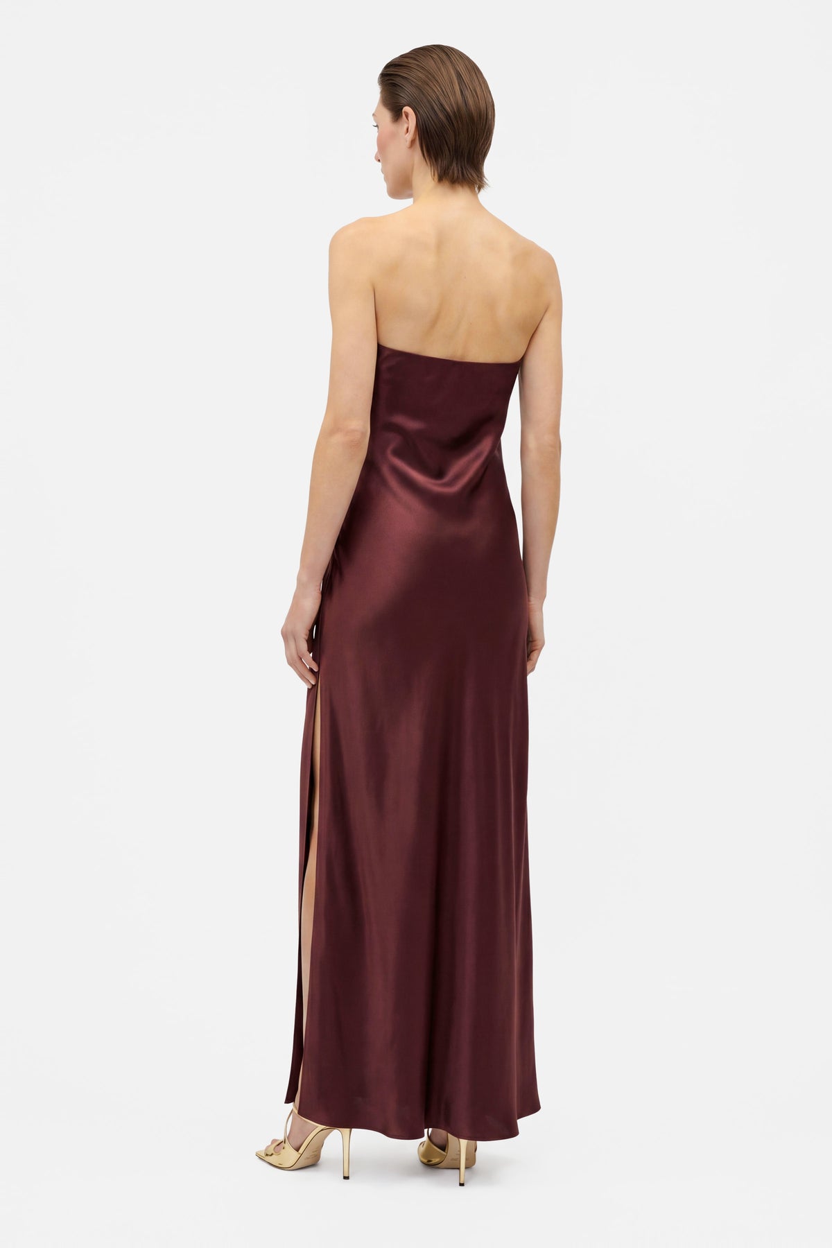Back view of model wearing burgundy strapless slit gown, standing gracefully.