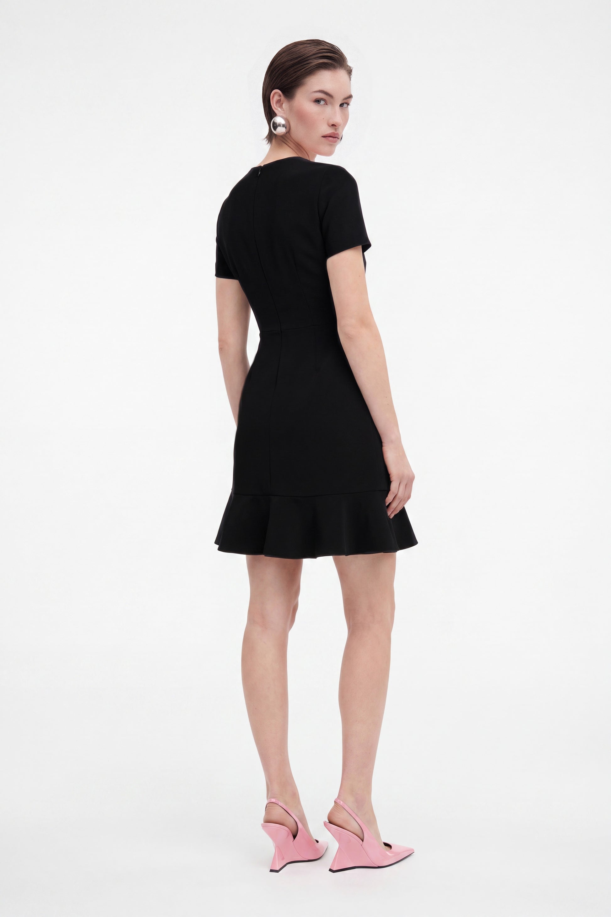 Model wearing black ruffle hem dress, back view, pink heels, sleek hairstyle.