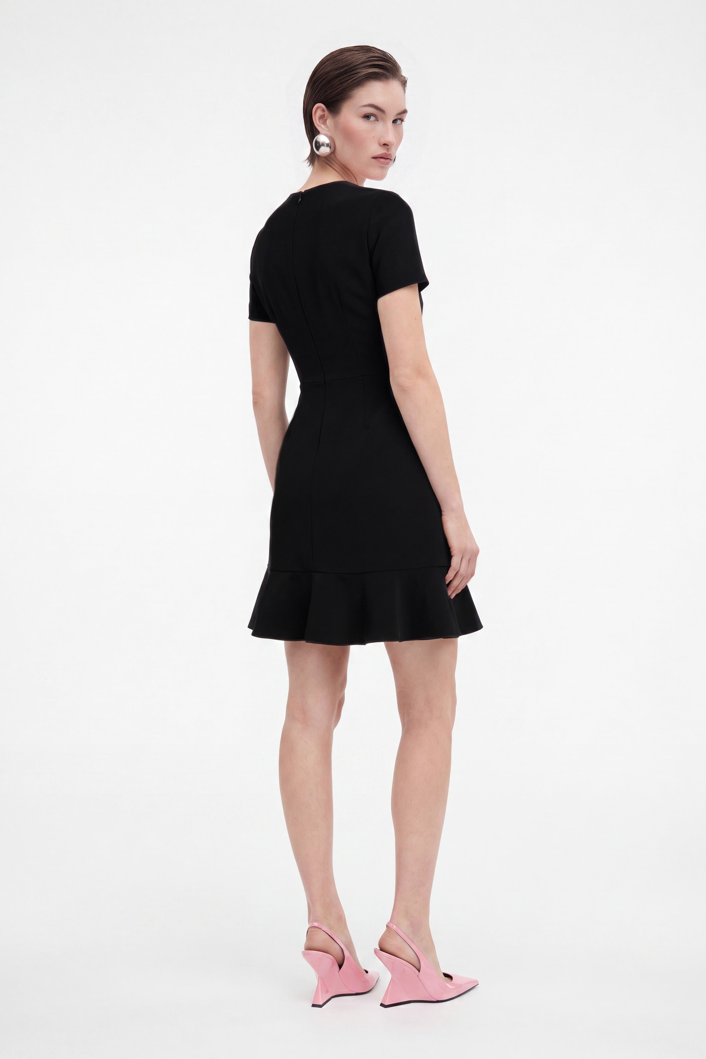 Model wearing black ruffle hem dress, back view, pink heels, sleek hairstyle.