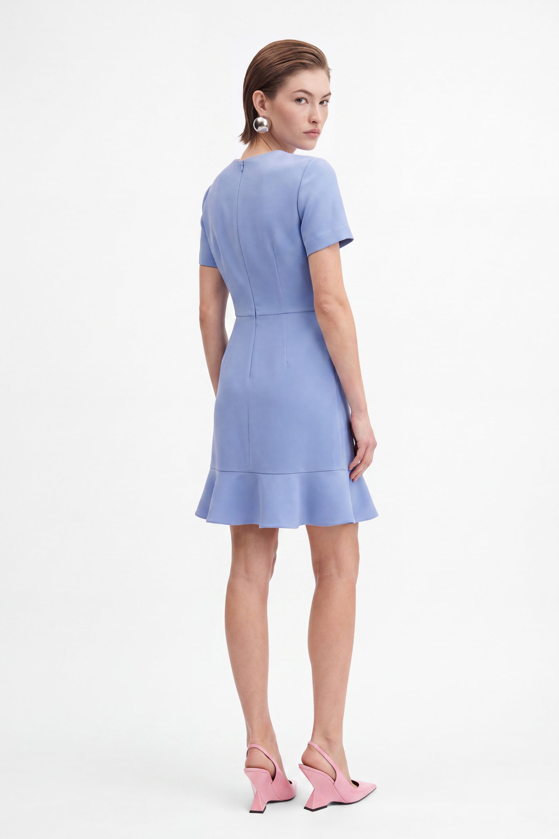 Back view of model wearing lavender flounce hem dress, pink heels, and silver earrings.