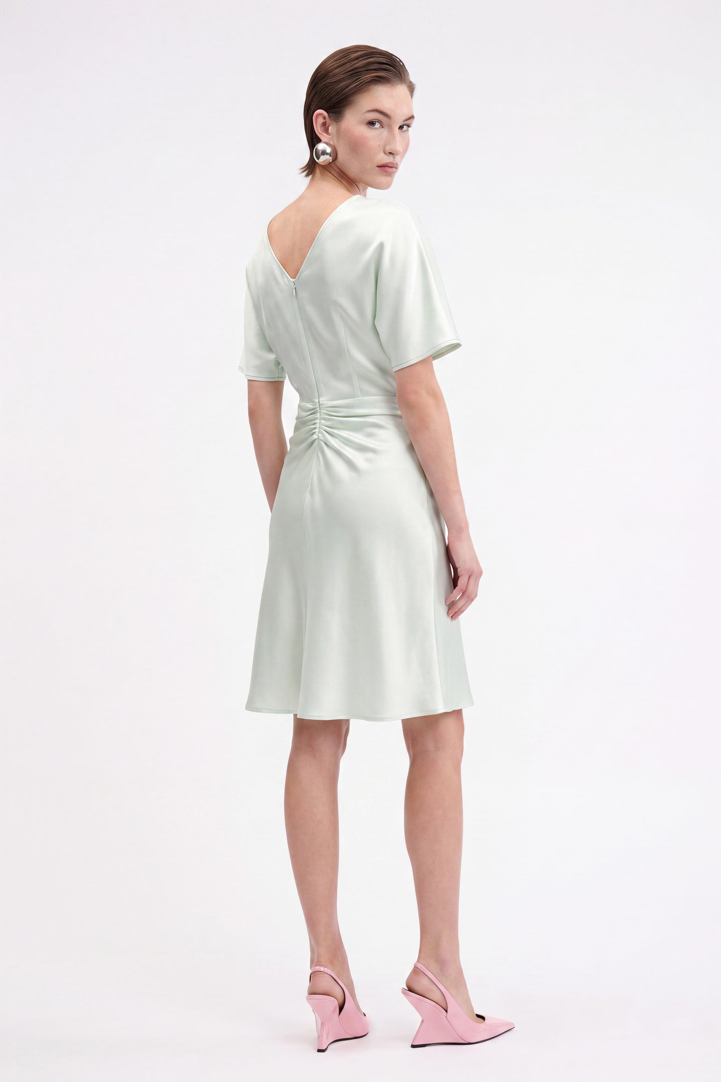 Model in mint ruched satin dress, back view, with pink heels and statement earrings.