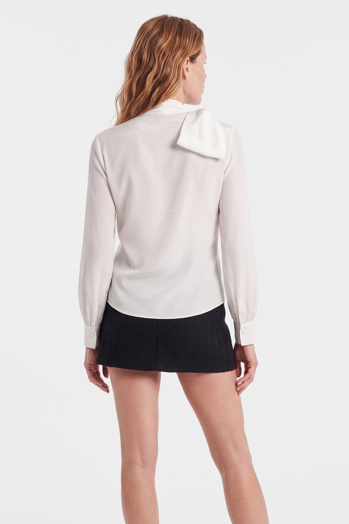 Model wearing ivory bow tie blouse and black mini skirt, back view.
