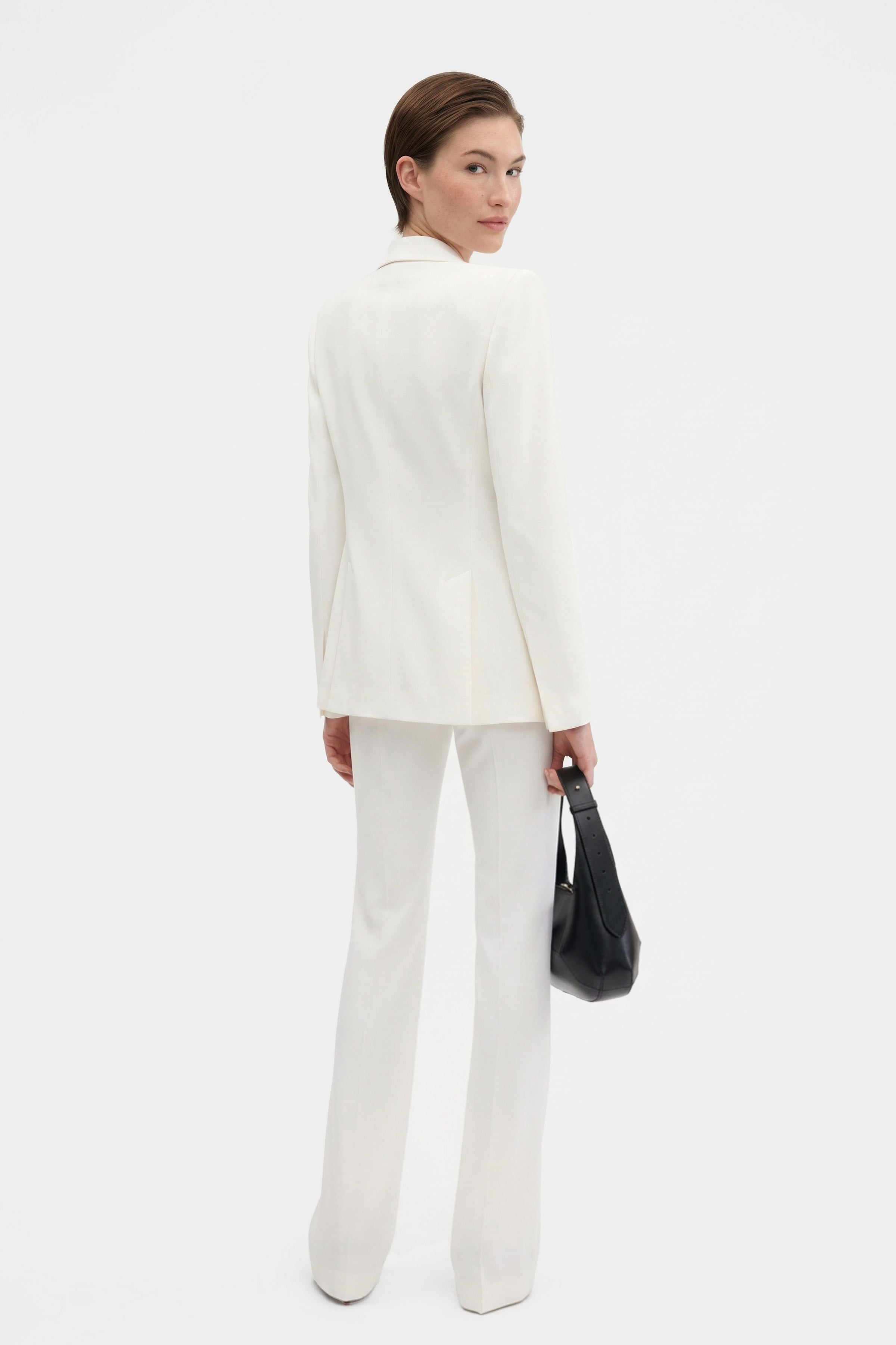 Model in ivory tailored blazer, back view, holding black bag, white background.