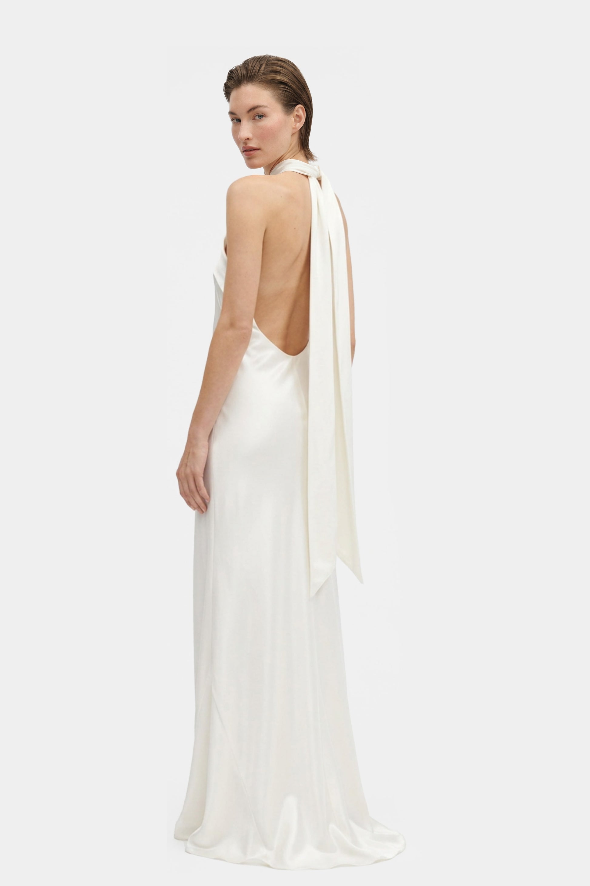 Model wearing elegant white silk halter gown, back view, flowy design, long tie detail.