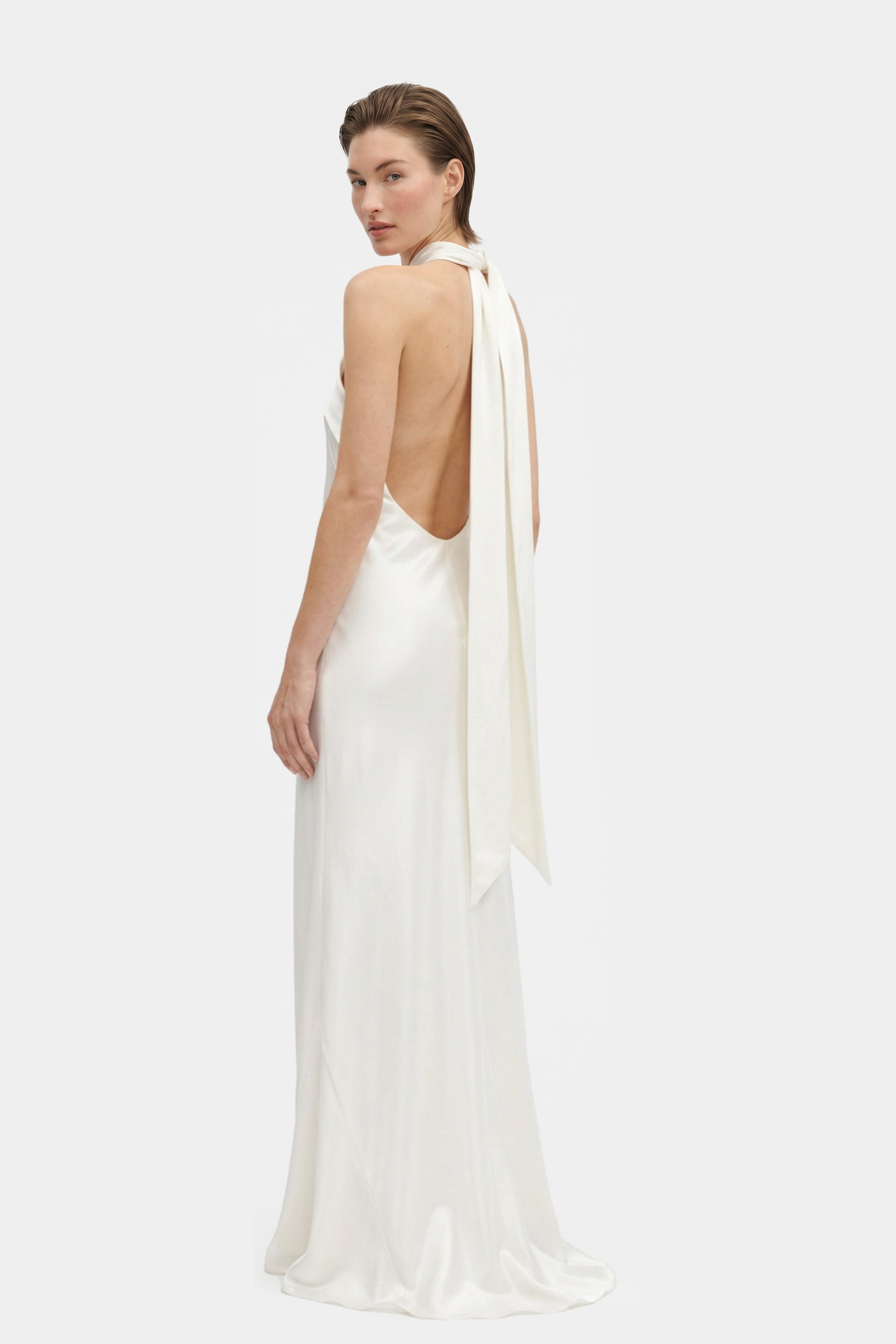 Model wearing elegant white silk halter gown, back view, flowy design, long tie detail.