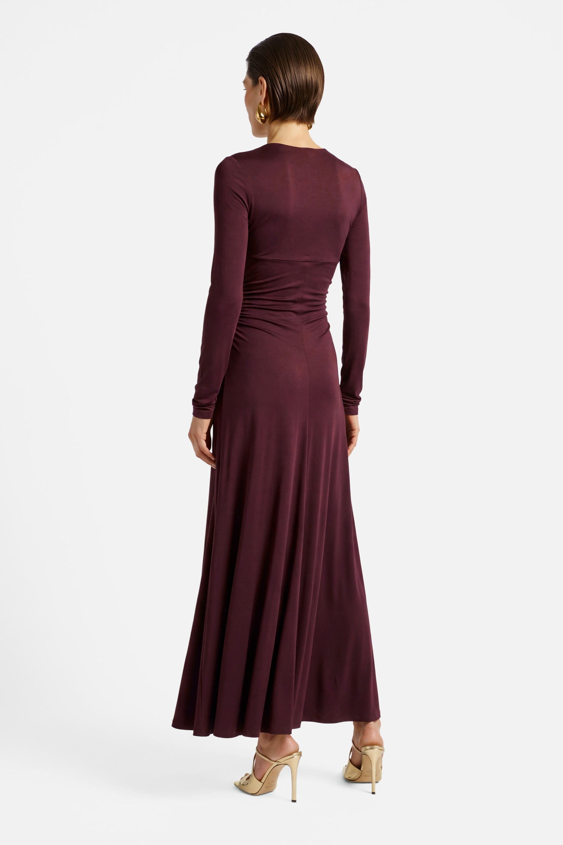 Model wearing burgundy ruched long sleeve dress, back view, gold high heels.