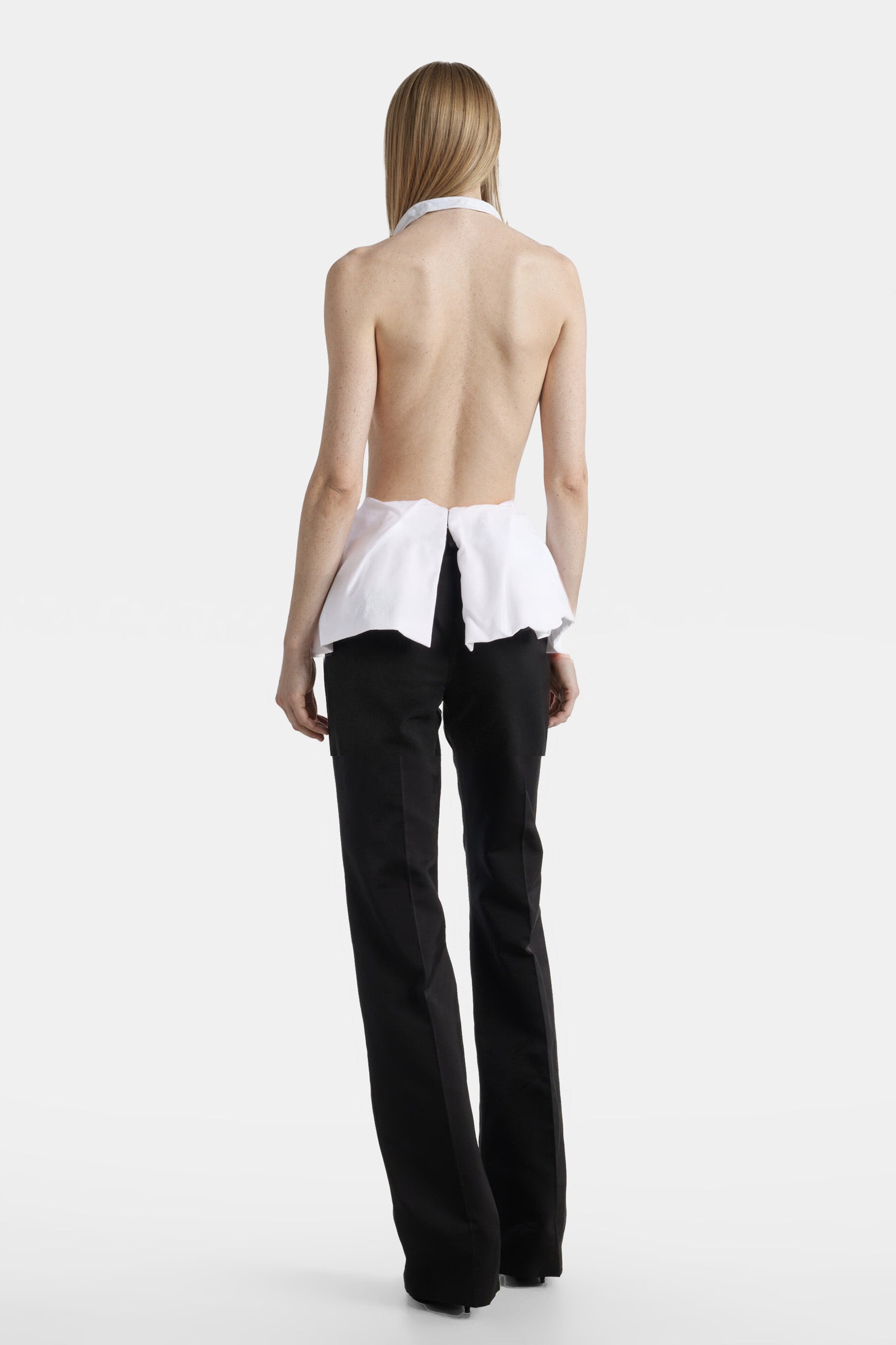 Back view of a white halter top with an open back and flared hem paired with black trousers.
