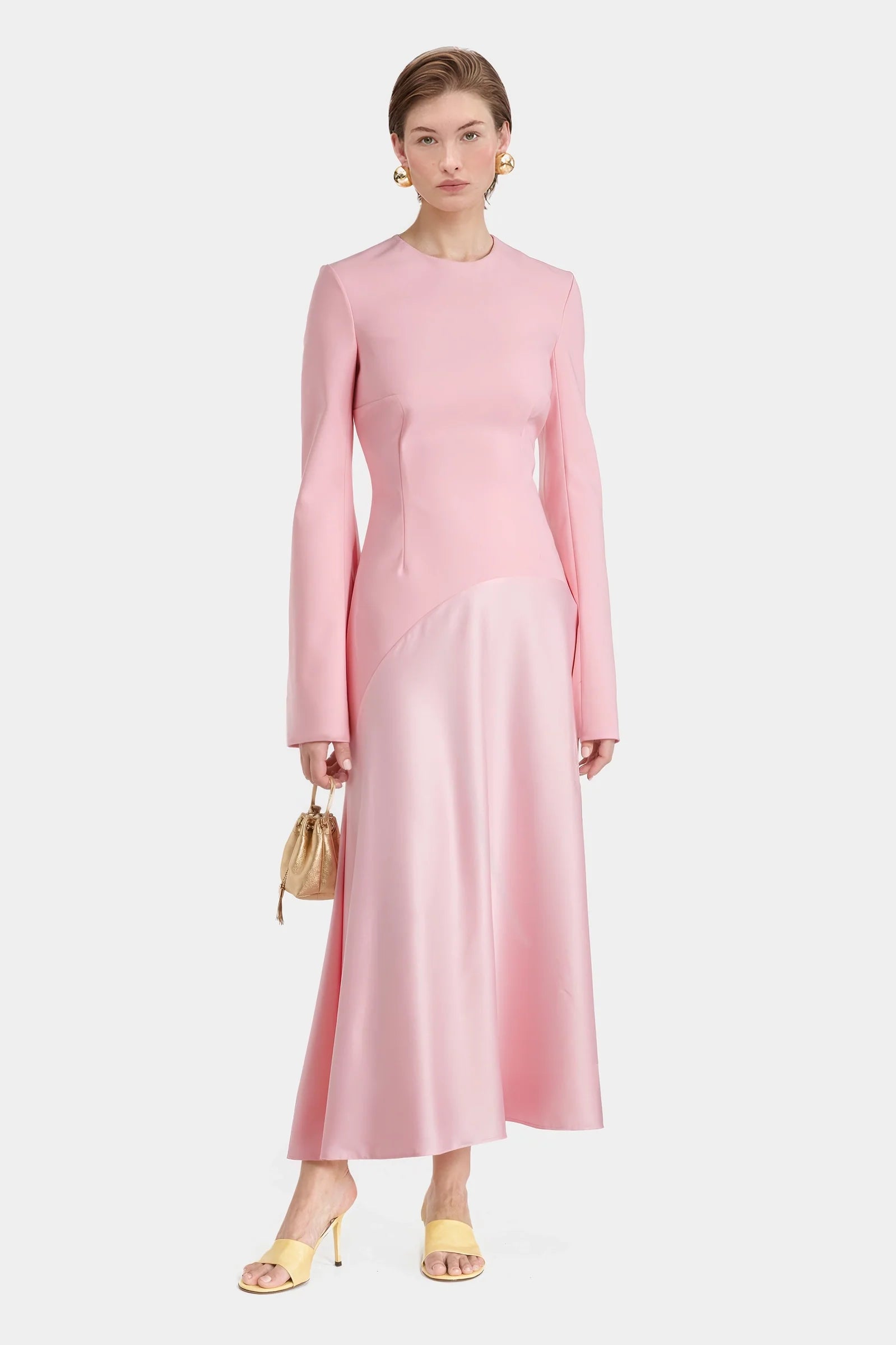 Model standing in a pink satin-crepe blend long dress with flared hem and long sleeves, holding a gold pouch bag.