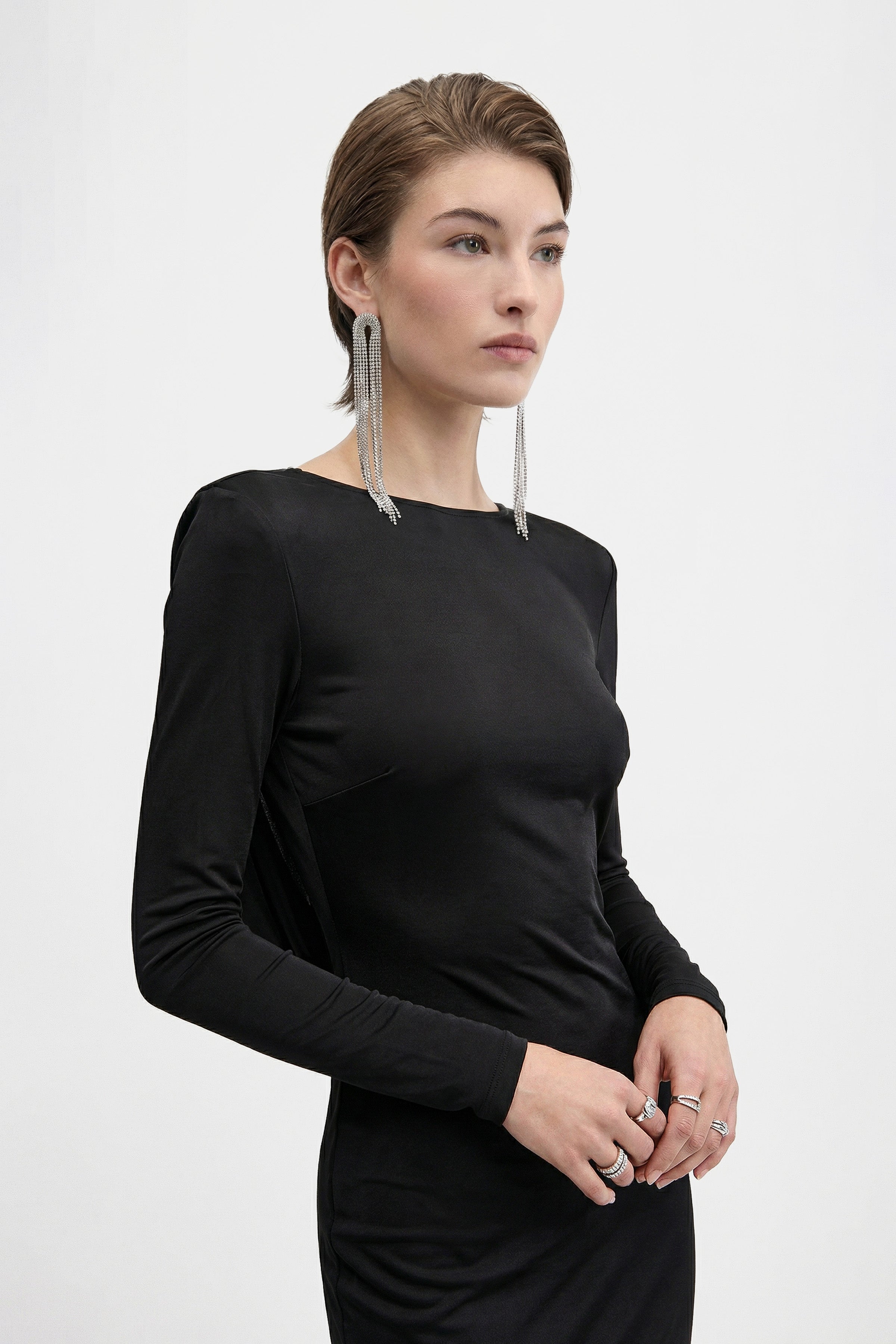 Three-quarter length front view of a woman wearing a minimalist black long-sleeved gown with a high neckline. She is styled with dramatic long crystal tassel earrings and several silver rings against a plain white background.