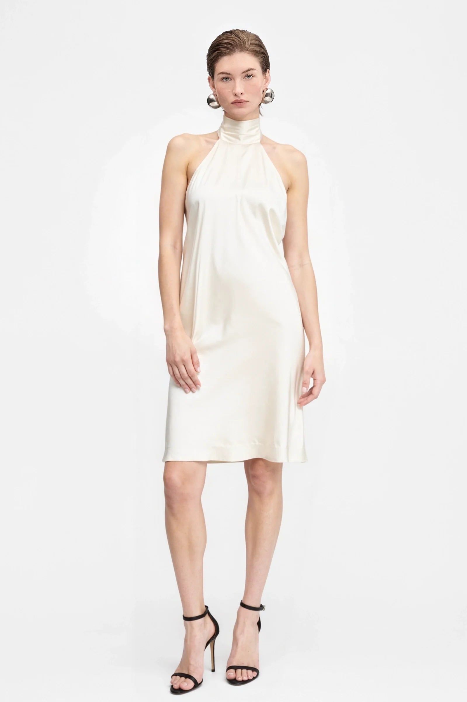 Model wearing cream satin mini dress with halter neck and open back, holding a small clutch.