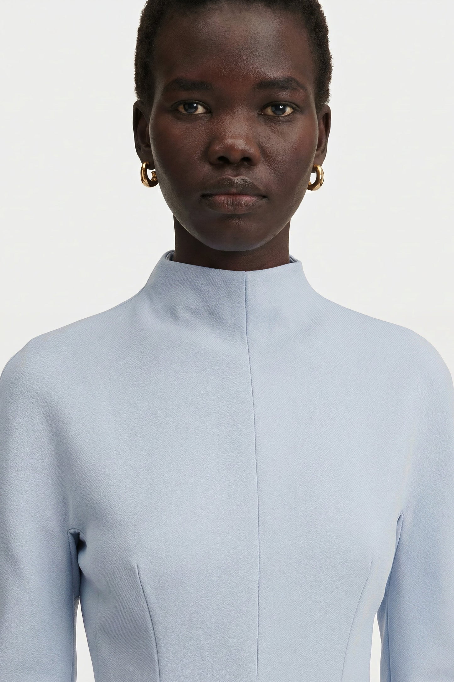 Close-up portrait showing the high mock neckline and structured shoulder detail of a light blue long-sleeve dress, accessorized with gold hoop earrings.