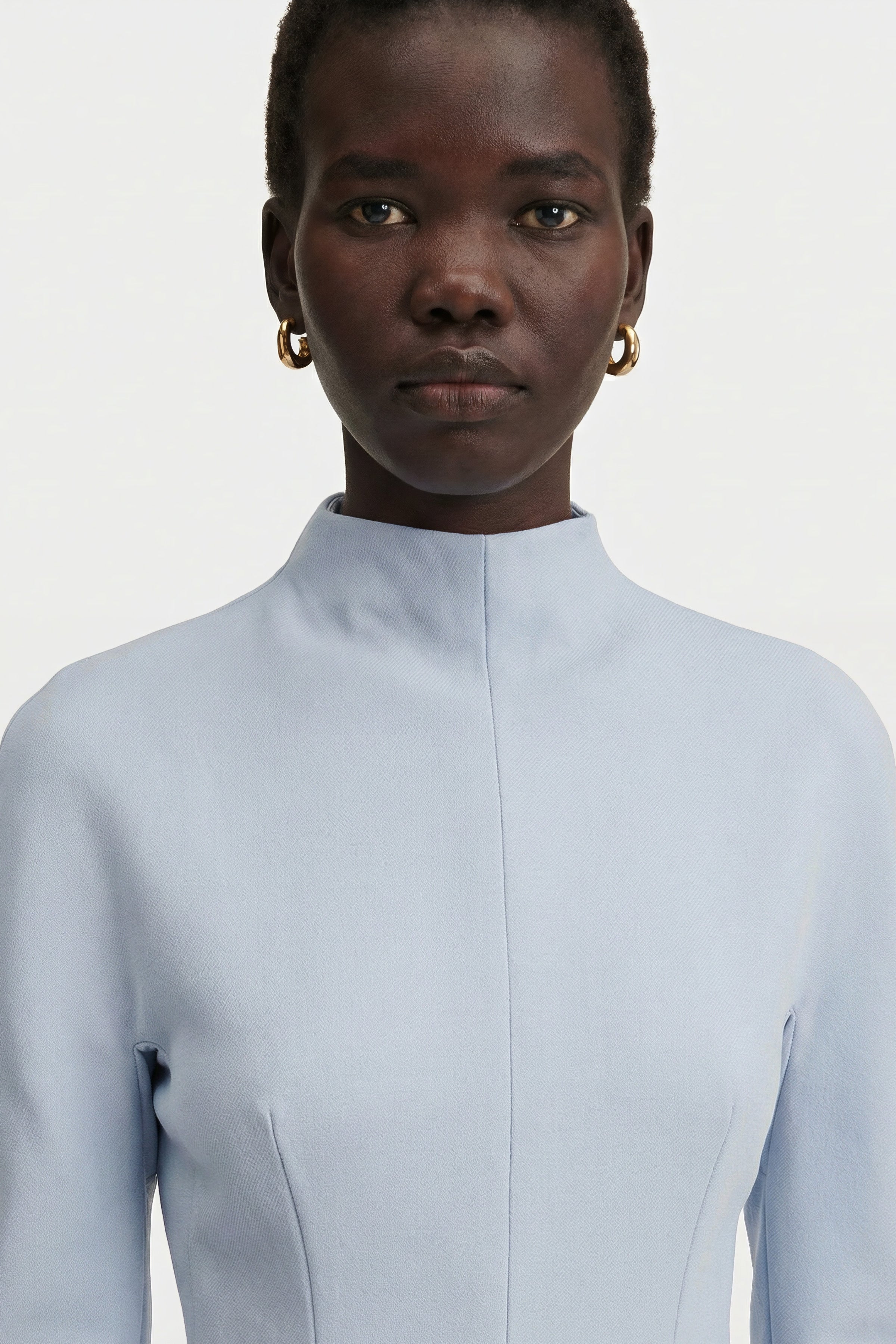 Close-up portrait showing the high mock neckline and structured shoulder detail of a light blue long-sleeve dress, accessorized with gold hoop earrings.